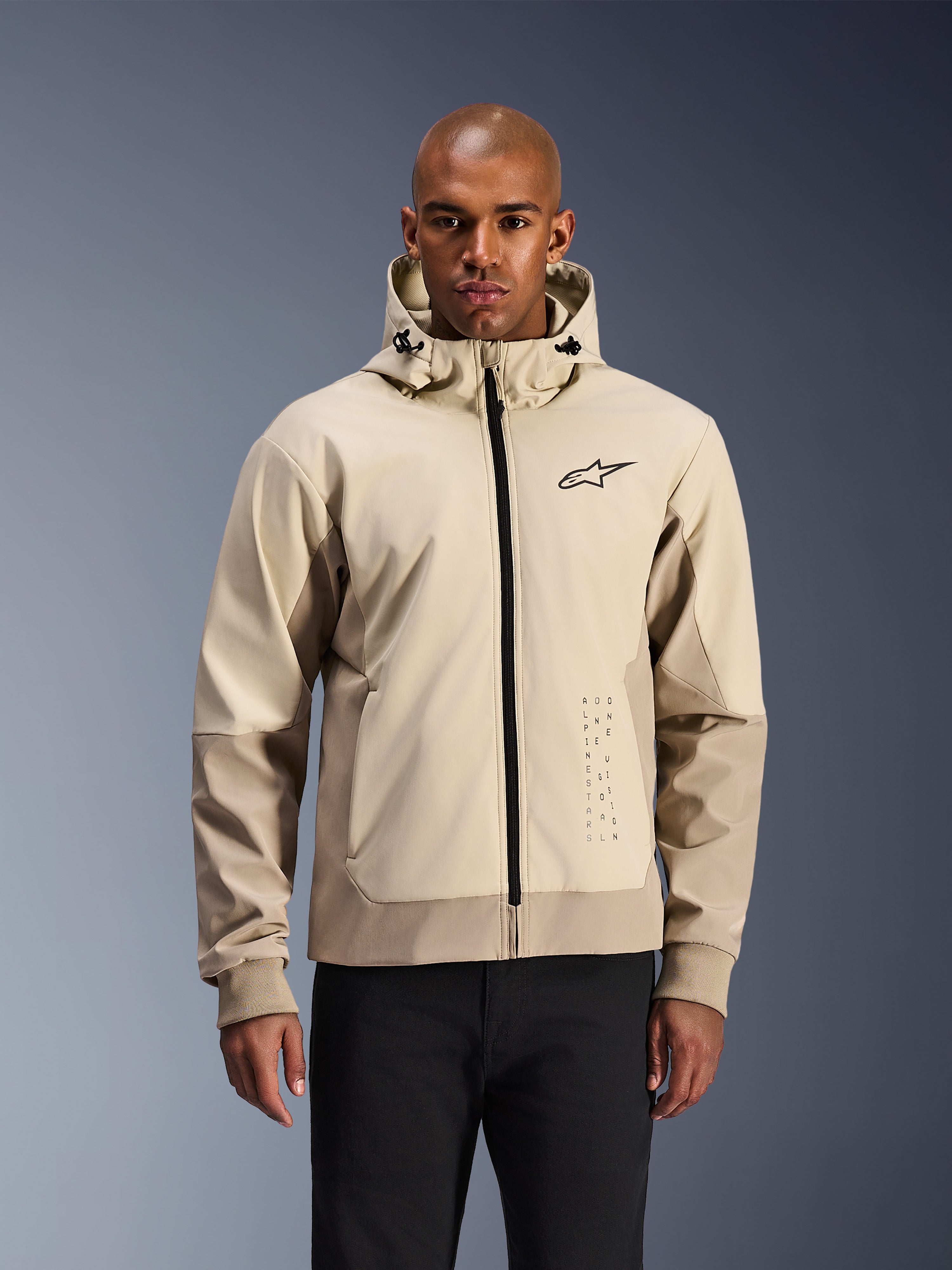 Alpinestars Radium Tech Hoodie in Mastic Aluminum, lichtbeige en tan stedelijke motorjas, zwarte rits aan de voorzijde en klein borstlogo, gedragen door een man in een zwarte broek, technische winddichte shell met eigentijdse streetwear-stijl
