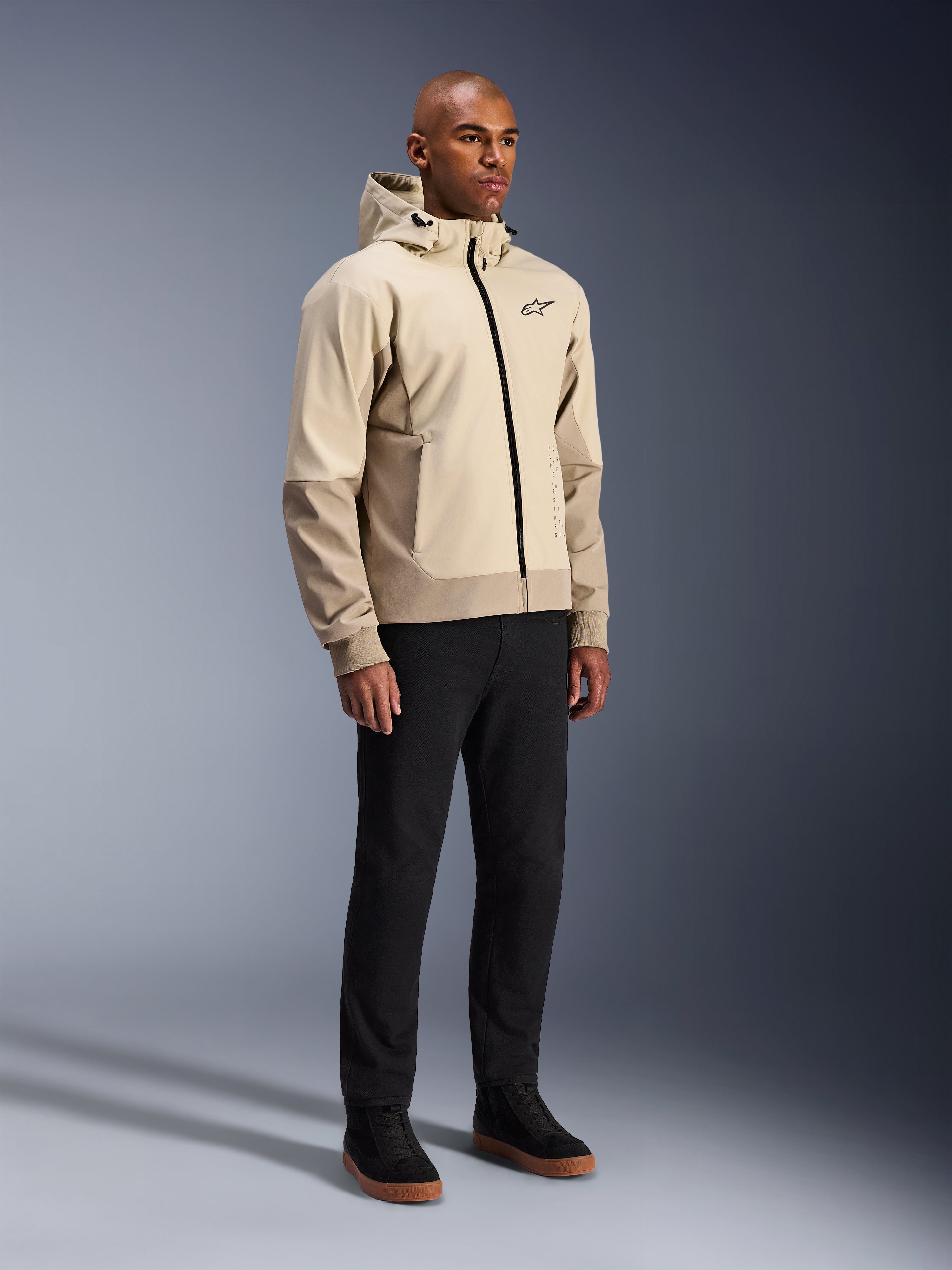 Alpinestars Radium Tech Hoodie, urban motorjas, Mastic Aluminum lichtbeige, gedragen met zwarte broek en zwarte hoge sneakers, voorzien van een technische winddichte buitenlaag, ritssluiting aan de voorzijde en afneembare capuchon, modern streetwear-design voor stedelijk rijden