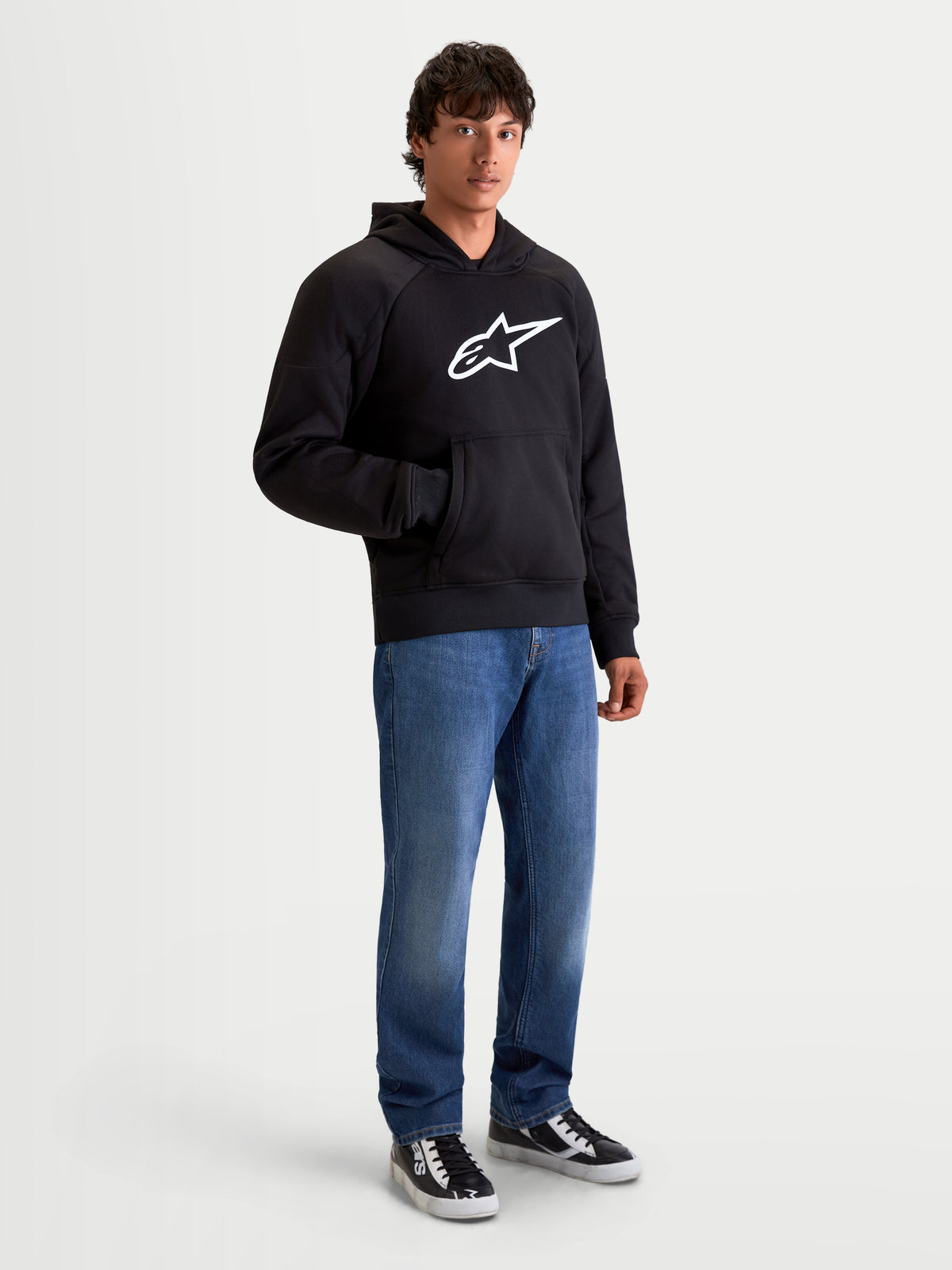 Alpinestars Ageless Hoodie, urban motorhoodie voor heren in zwart, vooraanzicht op model met casual spijkerbroek, voorzien van een prominent wit Astar-logo op de borst en interne slijtvaste verstevigingen
