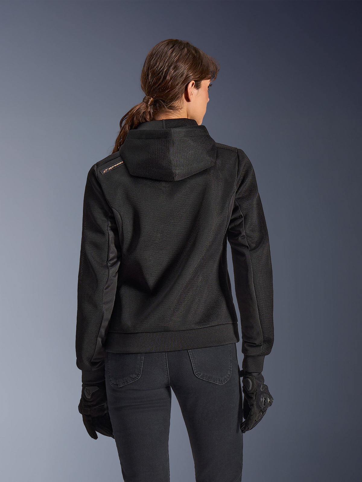 Alpinestars Stella Chrome Superairflow Hoodie, achteraanzicht van een vrouw die een zwarte technische 3D mesh motorhoodie draagt, voorzien van een technisch ontworpen capuchon en koperkleurig logo op de schouder, gedragen met zwarte motorhandschoenen en broek.