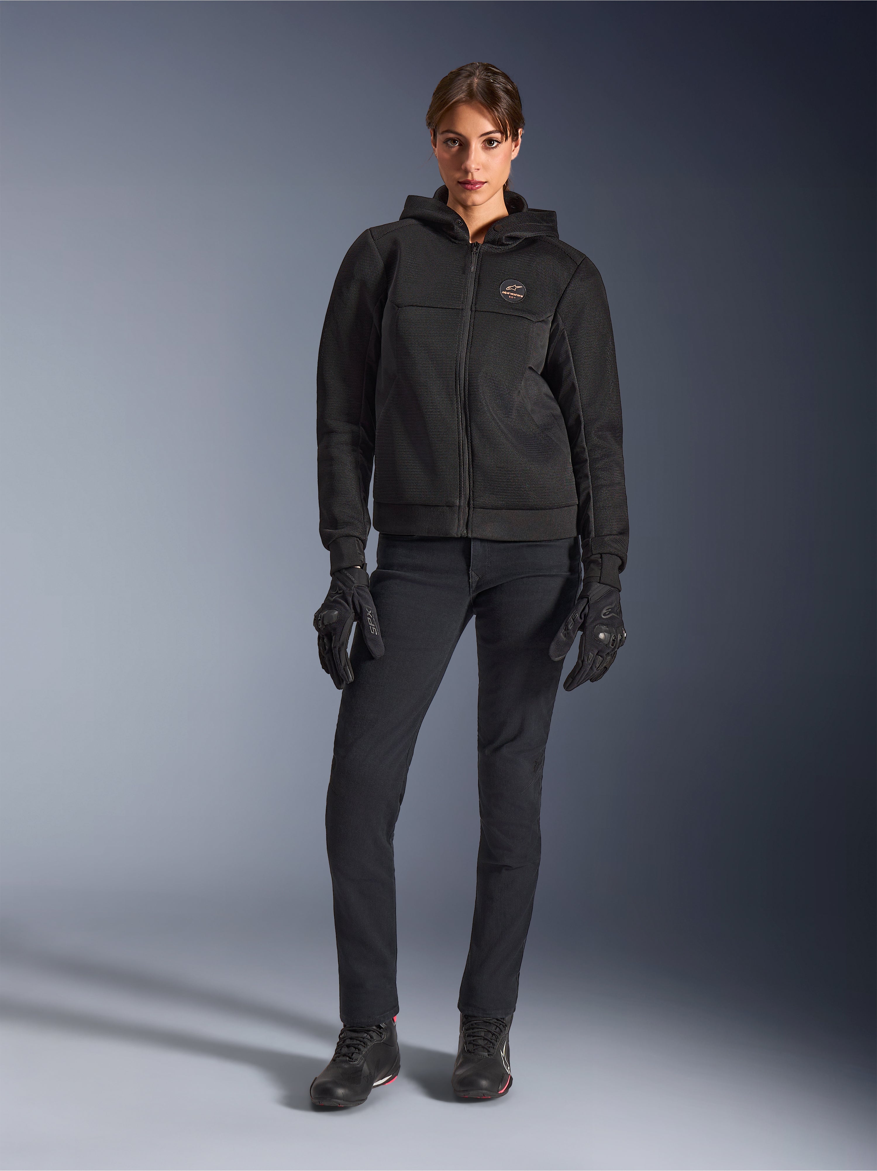 Female model wearing the Alpinestars Stella Chrome Superairflow Hoodie in Black, paired with black technical riding pants and gloves, featuring a 3D mesh textile construction and copper-toned chest logo, urban sport motorcycle apparel for women.