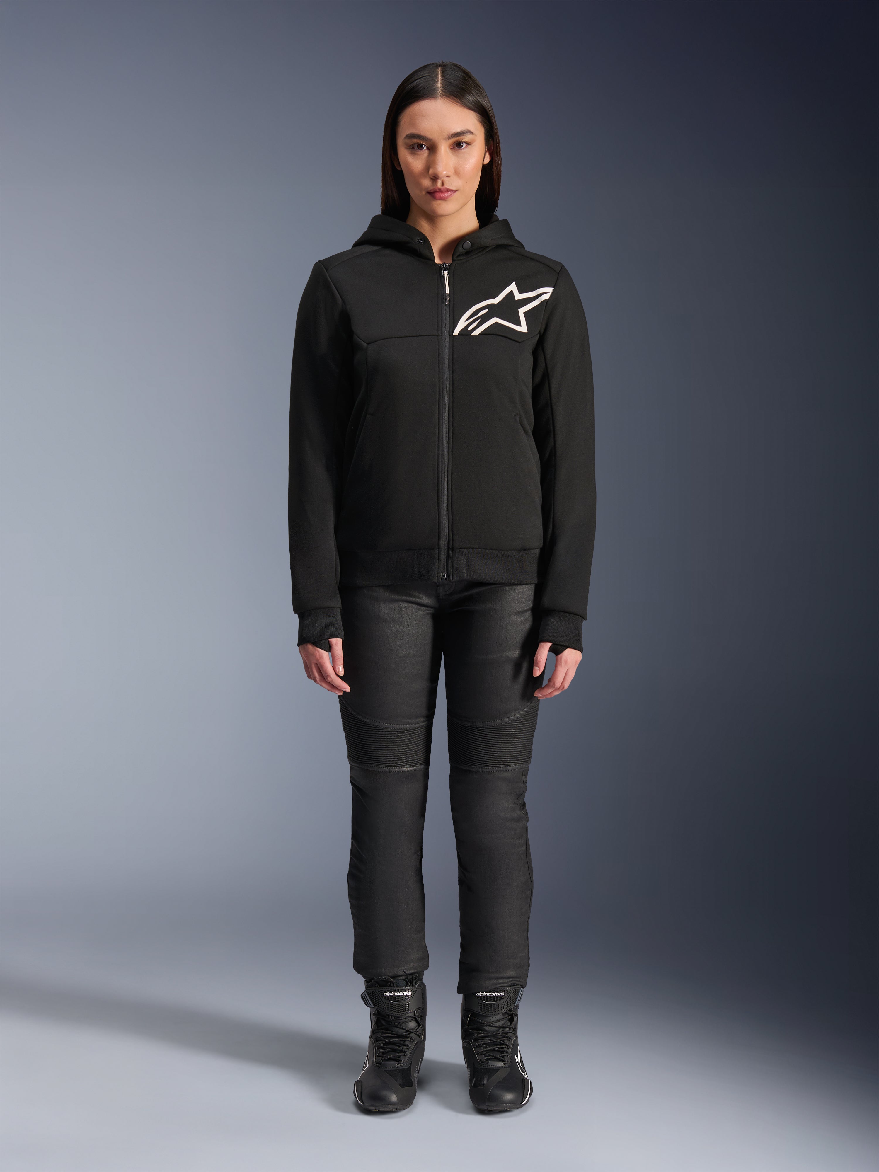 Woman wearing Alpinestars Stella Chrome V2 Sport Hoodie, Black with white Astars logo on the chest, paired with black technical riding pants and boots, urban motorcycle apparel design.