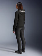 Woman wearing Alpinestars Stella Chrome V2 Sport Hoodie, rear view, black motorcycle hoodie with white Alpinestars logo on the hood, paired with black technical riding pants and black motorcycle shoes, casual urban commuting gear