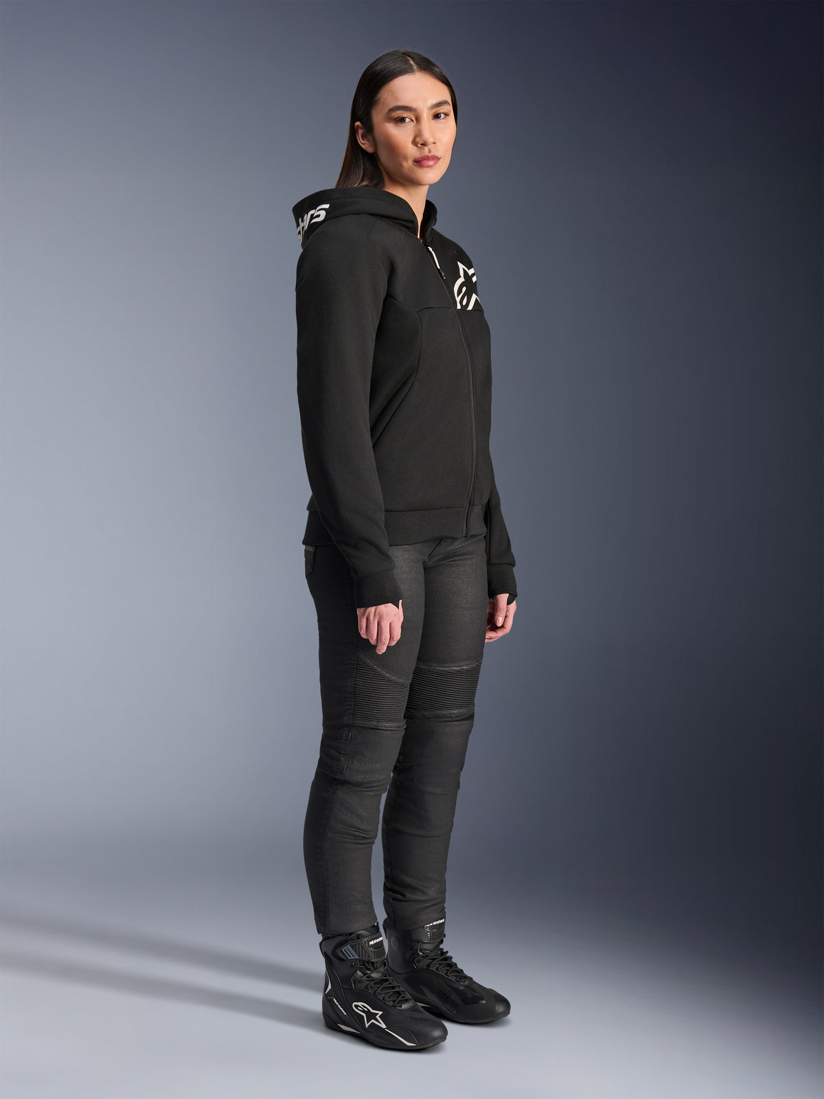 Vrouw die de Alpinestars Stella Chrome V2 Sport Hoodie draagt in het zwart, voorzien van witte Alpinestars branding, gecombineerd met een zwarte motorbroek en laarzen, urban motorkleding voor dames met een casual sportieve pasvorm