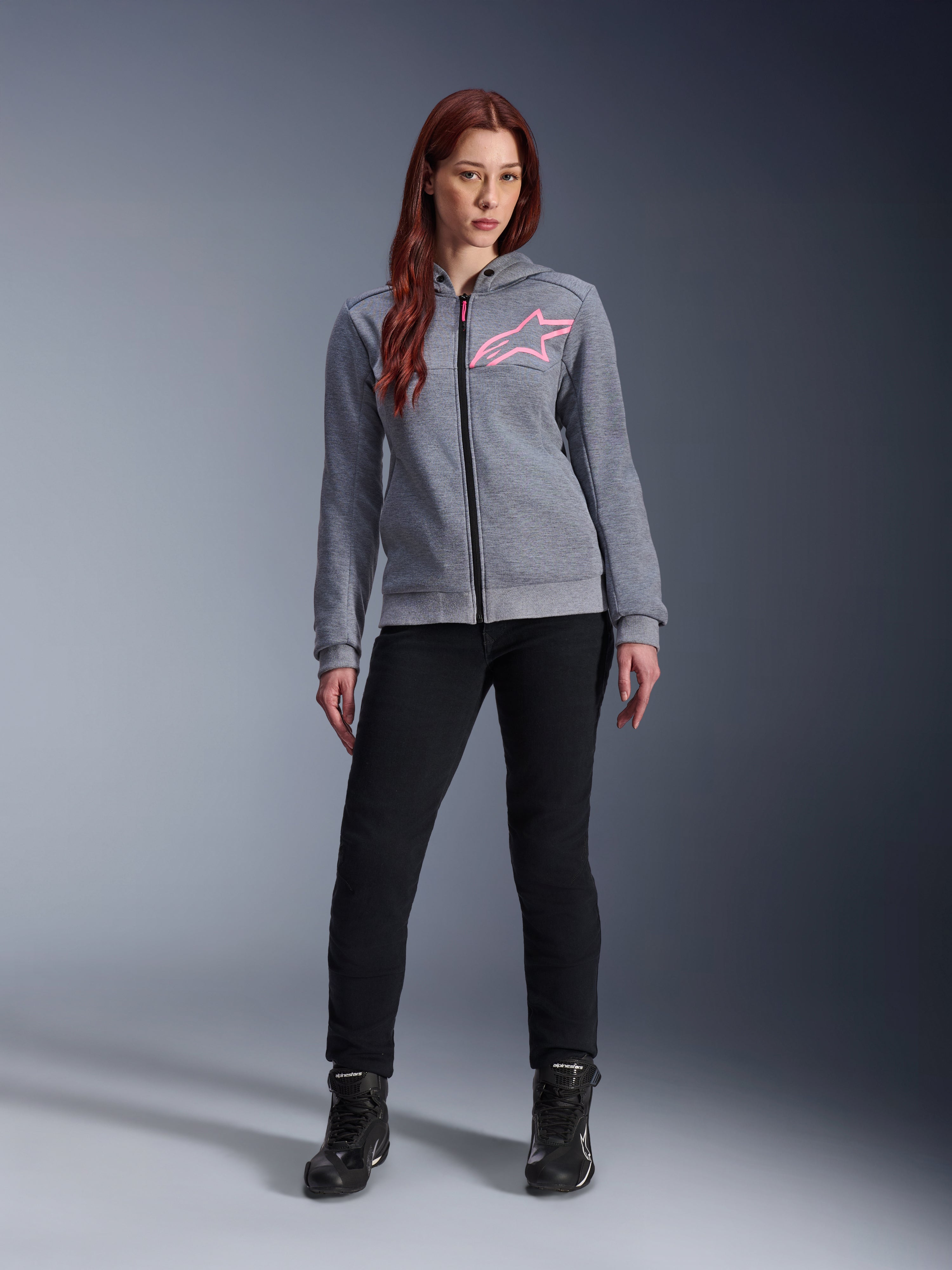 Alpinestars Stella Chrome V2 Sport Hoodie, Gray Heather Pink Fluo, gedragen door een vrouw met zwarte broek en laarzen, met een roze Alpinestars-sterlogo op de borst, urban motorkleding met volledige ritssluiting, lichtgewicht tech fleece met een casual atletische pasvorm.