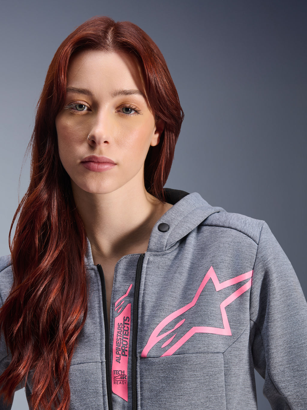 Vrouw draagt Alpinestars Stella Chrome V2 Sport Hoodie, Gray Heather Pink Fluo, close-up opname van het roze Alpinestars sterlogo en Tech-Air Ready branding, stedelijke motorkleding.