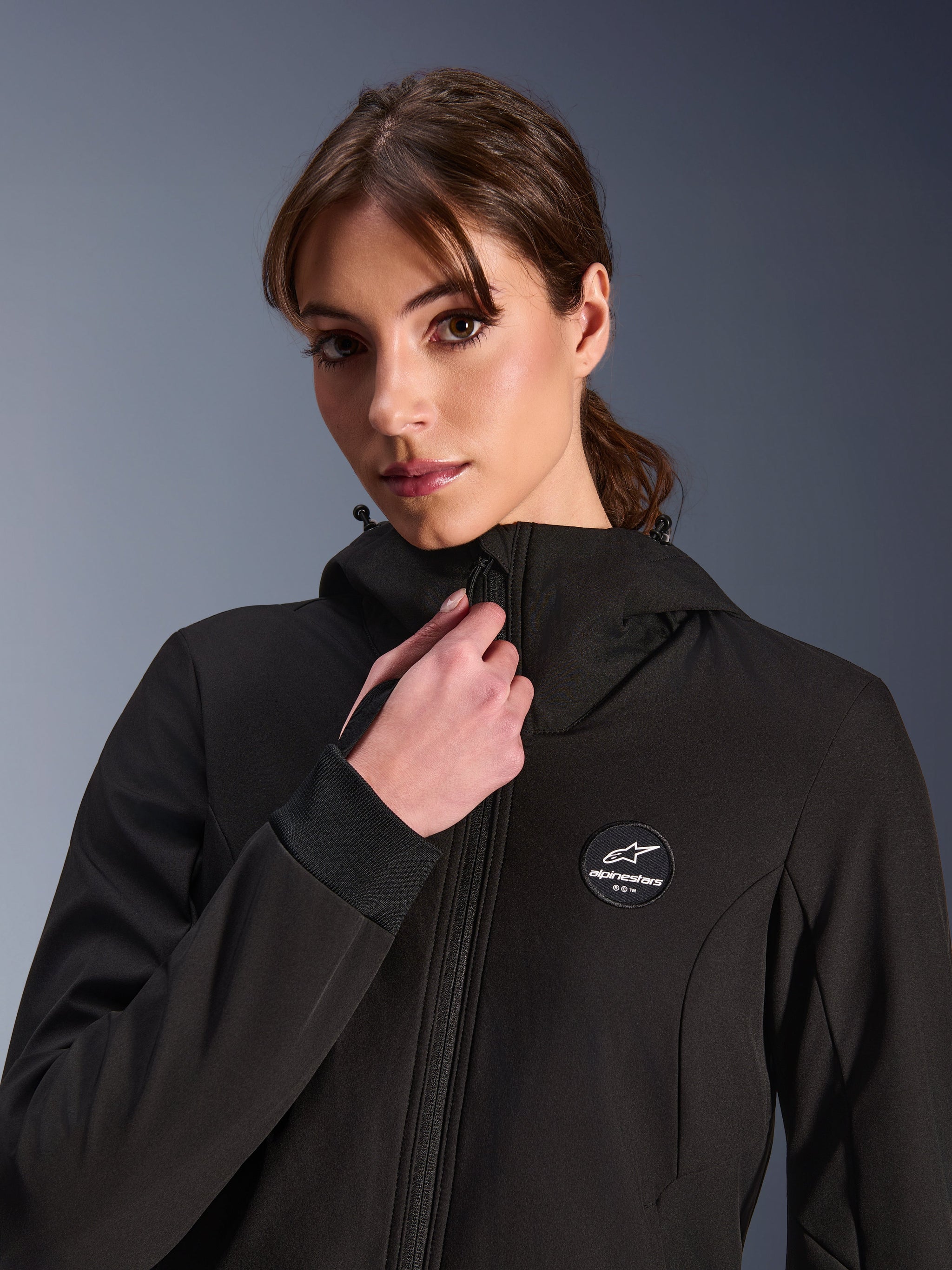Woman Stella Radium Tech Hoodie, Black, worn by a woman adjusting the front zipper, featuring circular Alpinestars logo patch on the chest, ribbed stretch cuffs, and windproof textile construction for urban riding