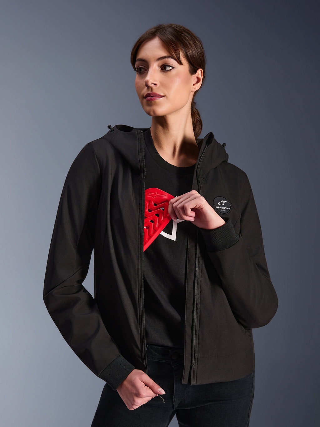 Alpinestars Woman Stella Radium Tech Hoodie, Black urban riding hoodie, female model demonstrating red Nucleon Flex Plus armor inside, lightweight 3-layer windproof textile shell with ribbed cuffs and chest logo patch, protective motorcycle apparel