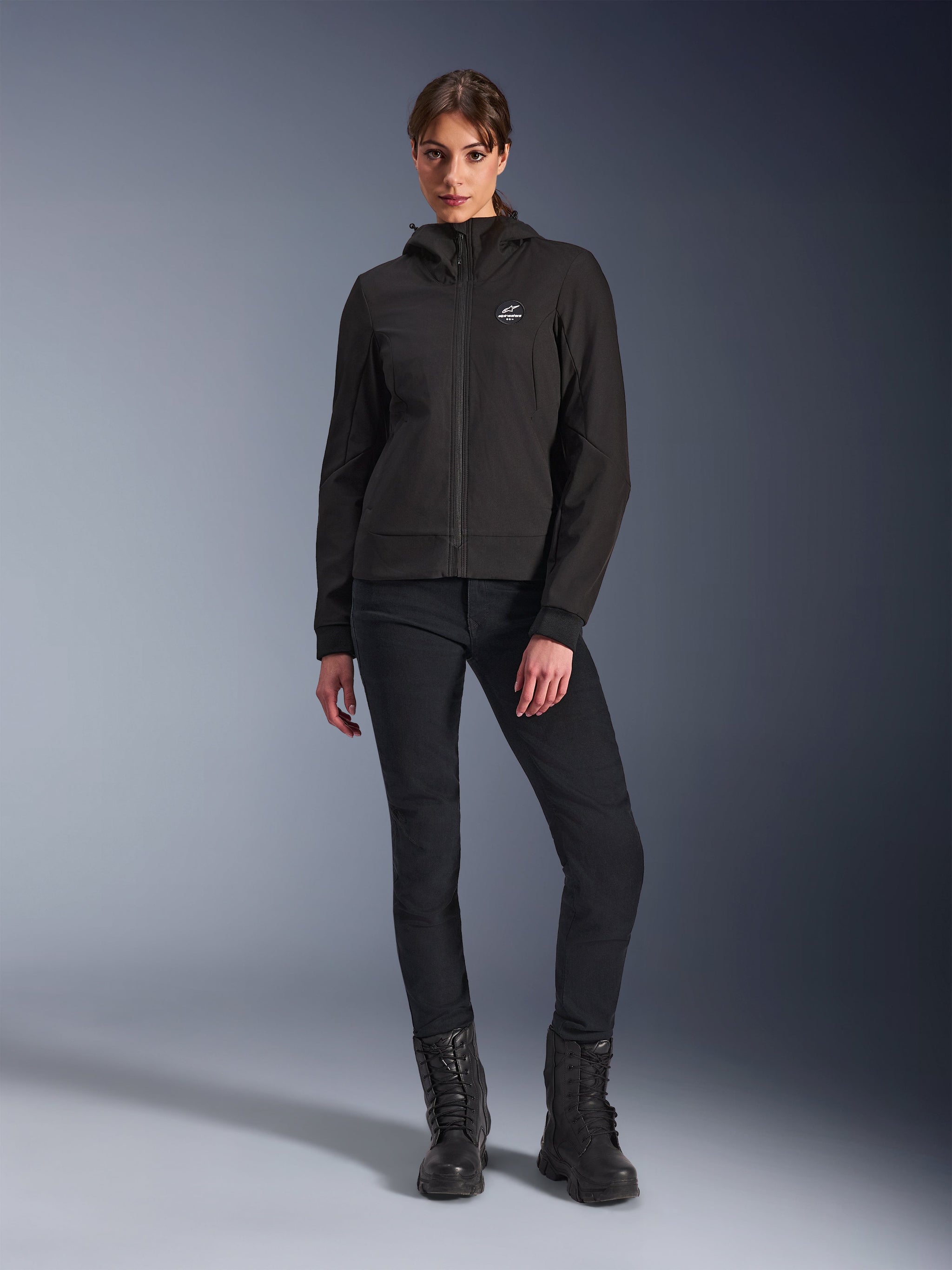 Alpinestars Stella Radium Tech Hoodie in Black, Women's urban motorcycle tech shell, worn with black slim-fit pants and black boots, featuring a small chest logo and hood, lightweight windproof construction for urban riding