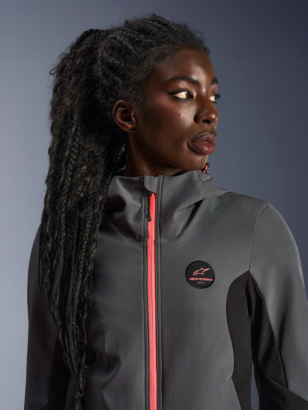 Vrouwelijk model draagt de Alpinestars Stella Radium Tech Hoodie in donkergrijs en zwart, met een fel koraalkleurige rits, rond logo op de borst en winddichte shell met hoge kraag, casual stedelijke motorkleding voor dames