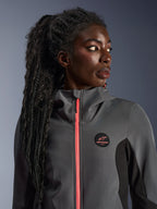 Vrouwelijk model draagt de Alpinestars Stella Radium Tech Hoodie in donkergrijs en zwart, met een fel koraalkleurige rits, rond logo op de borst en winddichte shell met hoge kraag, casual stedelijke motorkleding voor dames