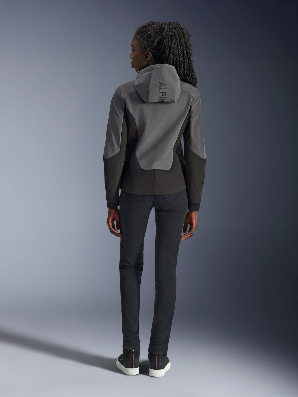 Alpinestars Stella Radium Tech Hoodie voor dames, donkergrijs en zwart, achteraanzicht met 'ALPIN'-branding op de capuchon, gecombineerd met een zwarte slim-fit broek en zwarte sneakers, stedelijke motorkleding