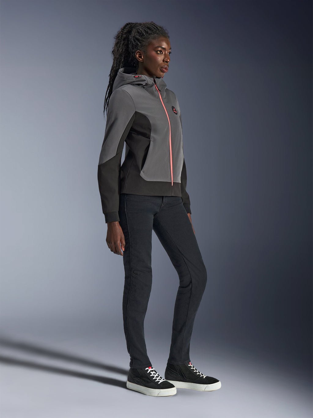 Alpinestars Stella Radium Tech Hoodie, urban motorhoodie, diepgrijs en zwart met een feloranje ritssluiting, gedragen met een zwarte broek en zwarte sneakers, vrouwspecifieke atletische pasvorm