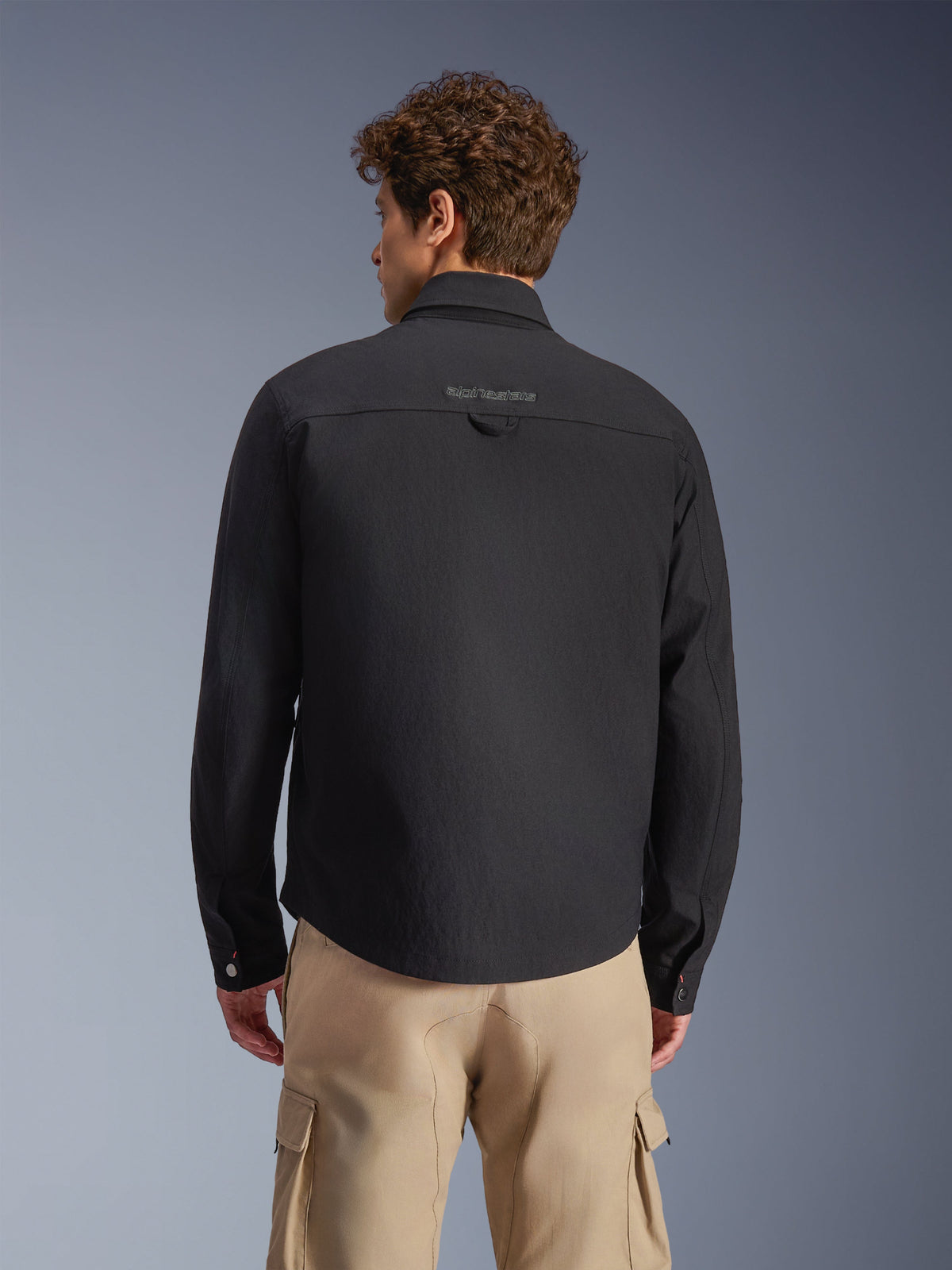 Alpinestars Bryon Water Resistant Overshirt, zwart, achteraanzicht gedragen door model, voorzien van geborduurd merklogo op het rugstuk en een functionele ophanglus, gestyled met een beige cargobroek, urban motorkleding met een klassieke workwear-esthetiek