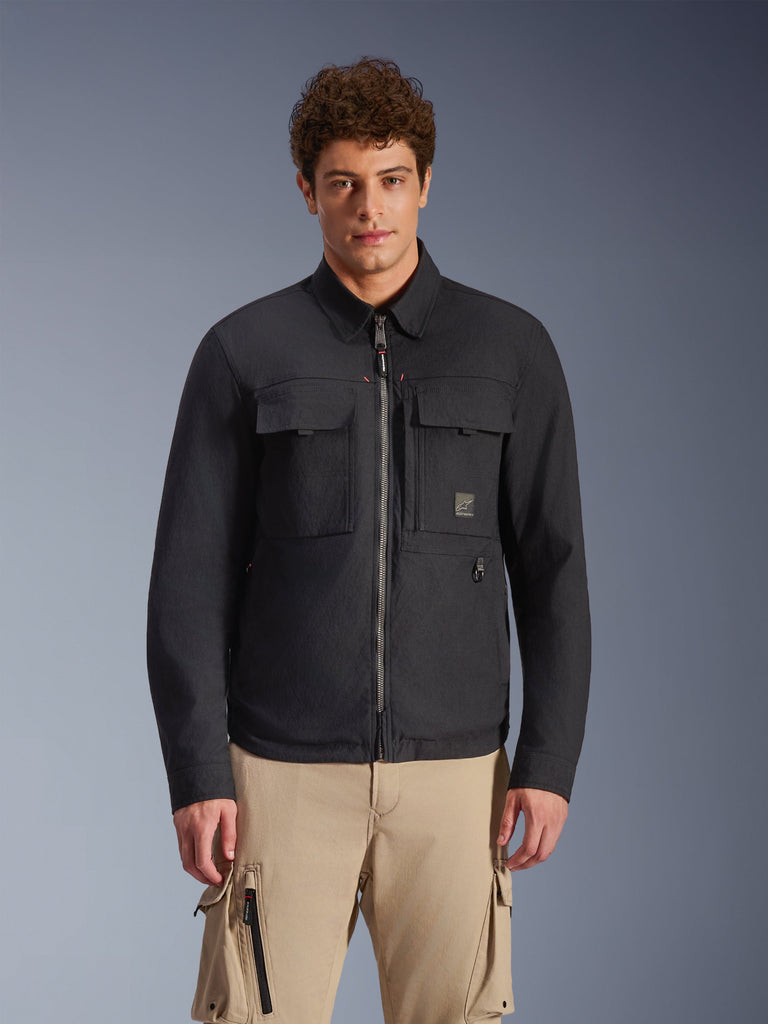 Alpinestars Bryon Water Resistant Overshirt, zwarte urban motorjas met op workwear geïnspireerde styling, voorzien van twee grote cargozakken op de borst en een volledige ritssluiting, gemaakt van gewaxt canvas, gedragen door een model met een beige cargobroek voor een eigentijdse utility-look.