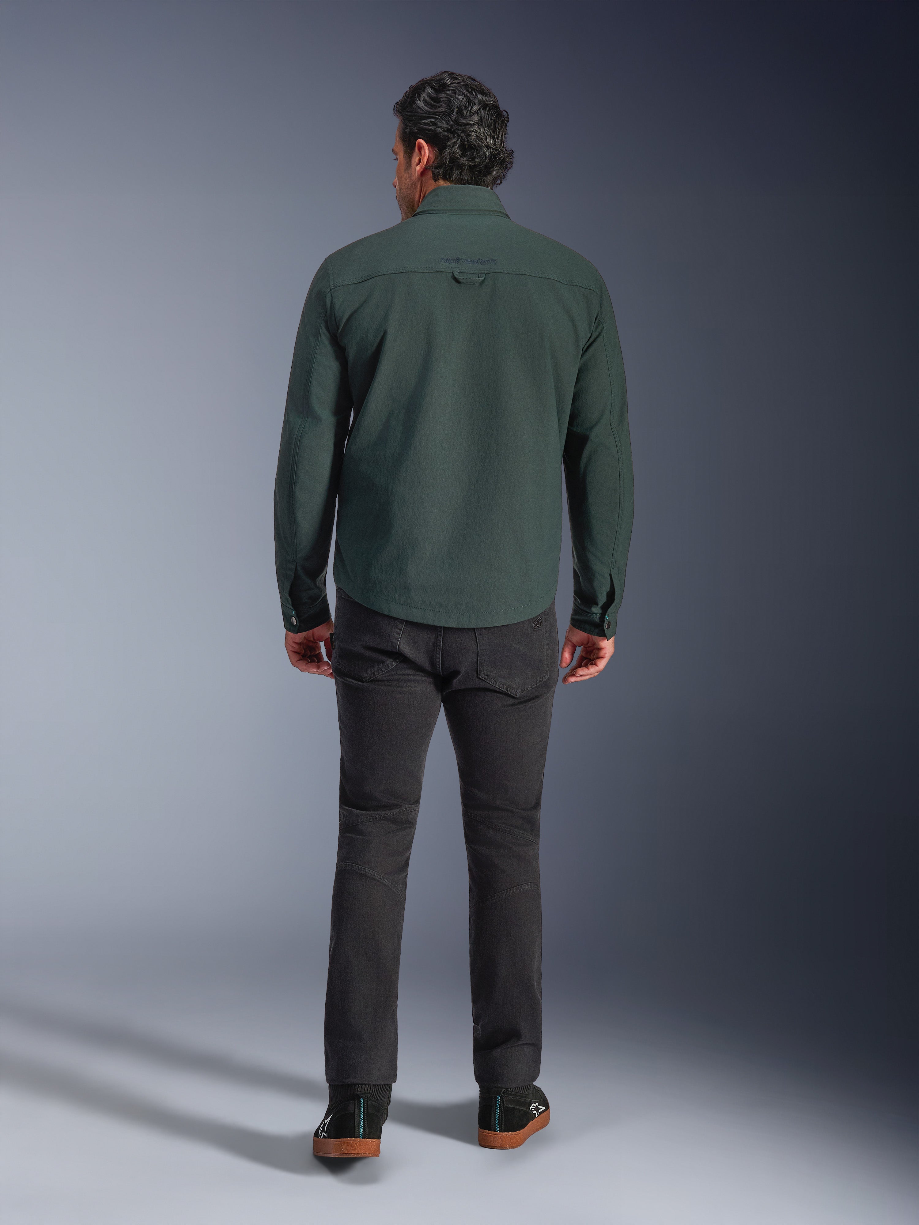 Alpinestars Bryon Water Resistant Overshirt in Dark Emerald, achteraanzicht op model, gecombineerd met een donkergrijze motorbroek en zwarte motorschoenen, klassiek op werkkleding geïnspireerd ontwerp met een iets langere achterkant voor verbeterde dekking tijdens het rijden.