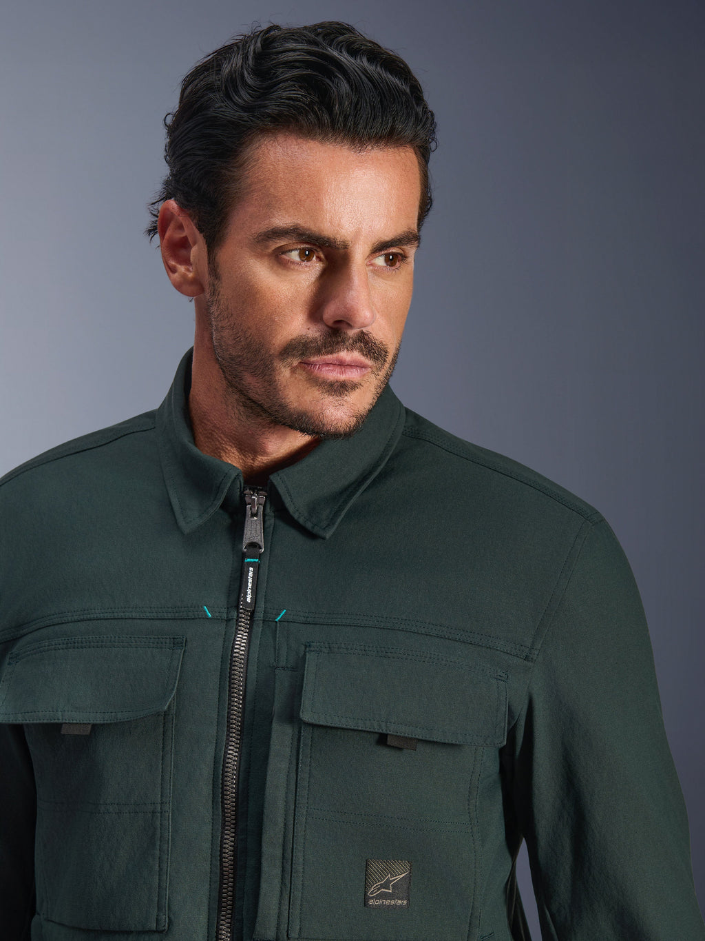 Alpinestars Bryon Water Resistant Overshirt in Dark Emerald, close-up portret van een man die de stedelijke motorjas draagt, gewaxte canvas textuur, voorrits met subtiele teal accenten, cargozakken op de borst en merklogo-patch.