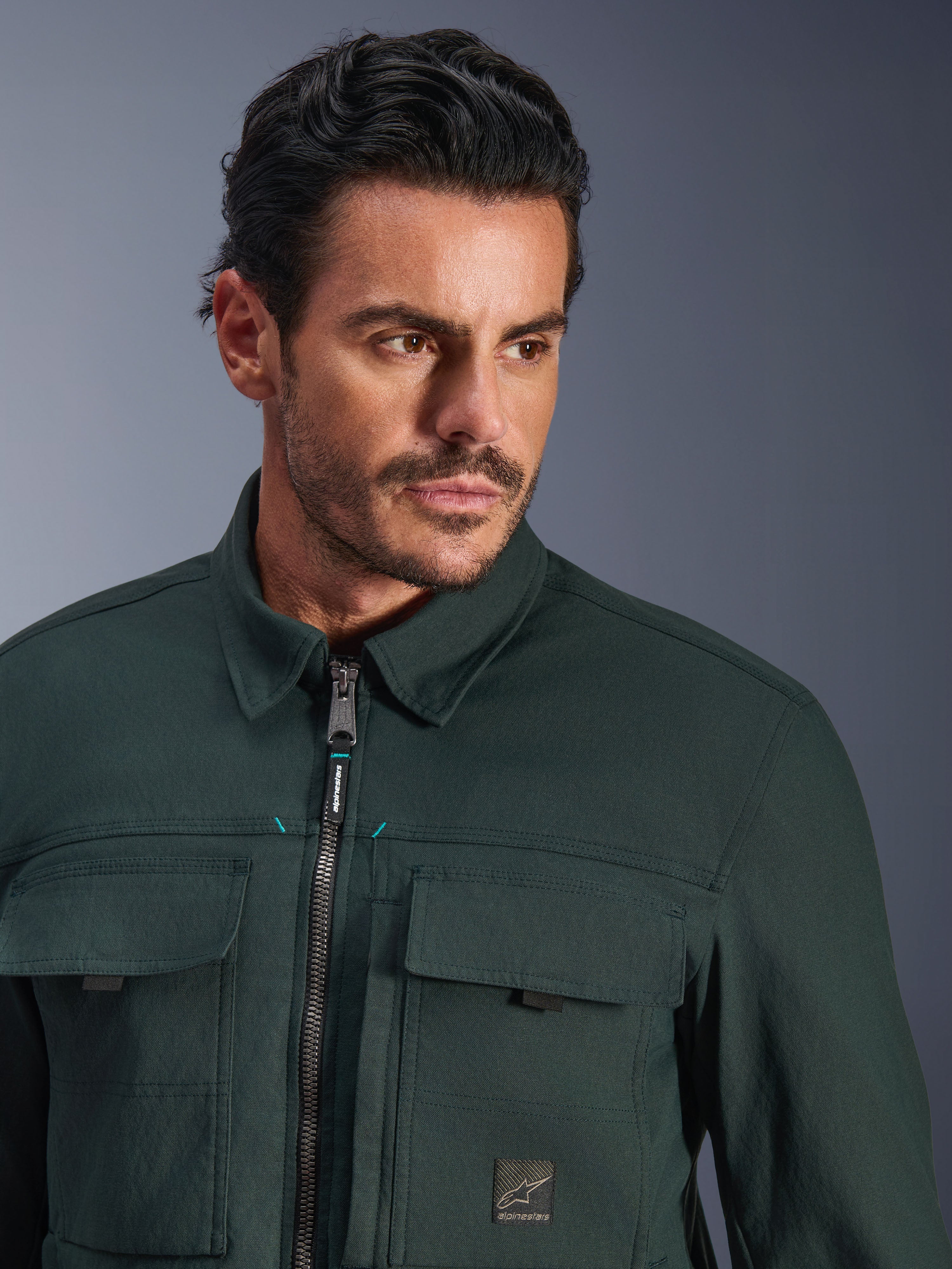 Alpinestars Bryon Water Resistant Overshirt in Dark Emerald, close-up portret van een man die de stedelijke motorjas draagt, gewaxte canvas textuur, voorrits met subtiele teal accenten, cargozakken op de borst en merklogo-patch.