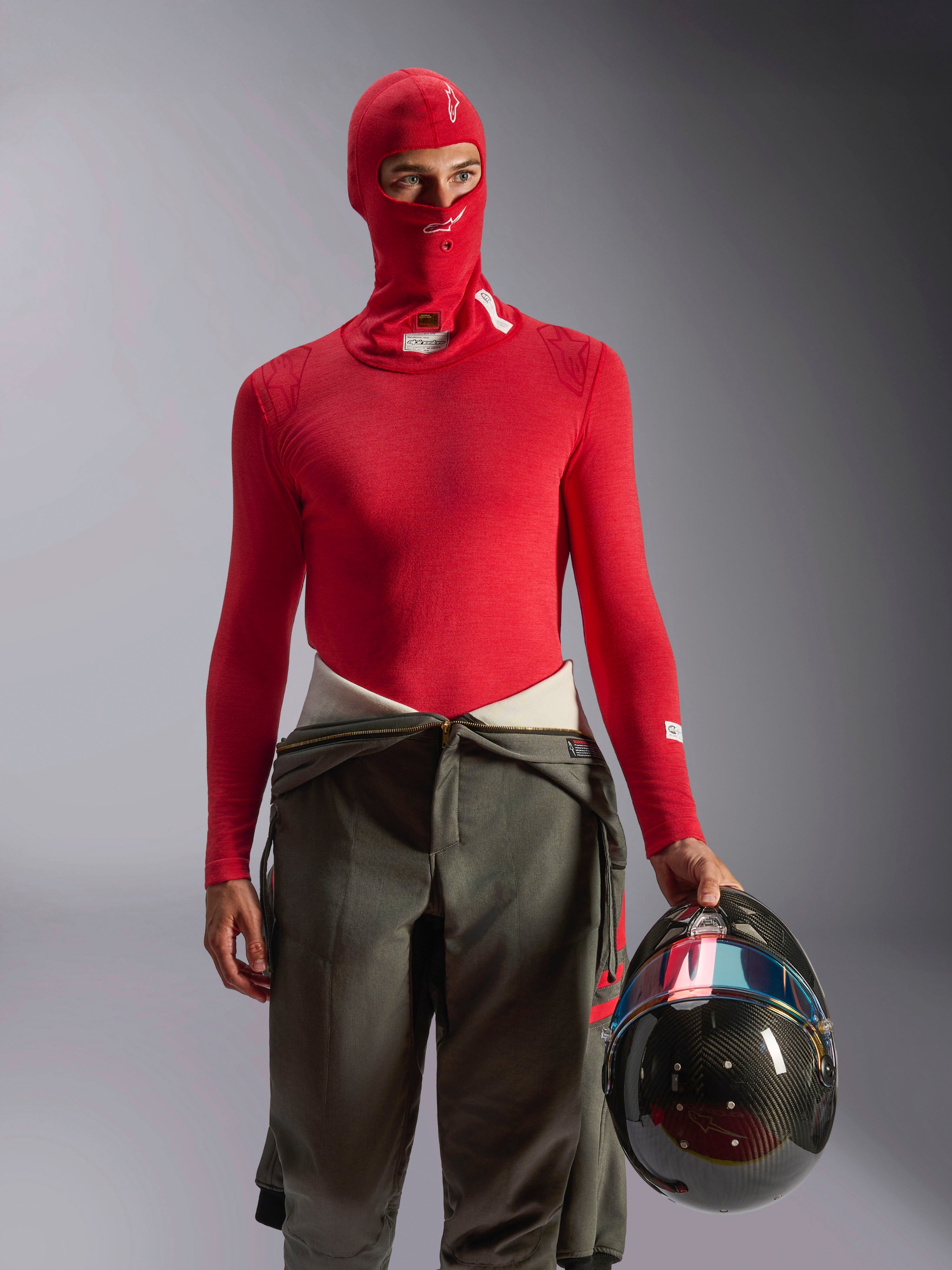 Alpinestars ZX Evo V3 Bivakmuts in rood, autorace-onderkleding gedragen door een coureur met bijpassend rood shirt met lange mouwen en olijfgroene racebroek, houdt een koolstofvezel helm vast, professionele brandwerende motorsportuitrusting
