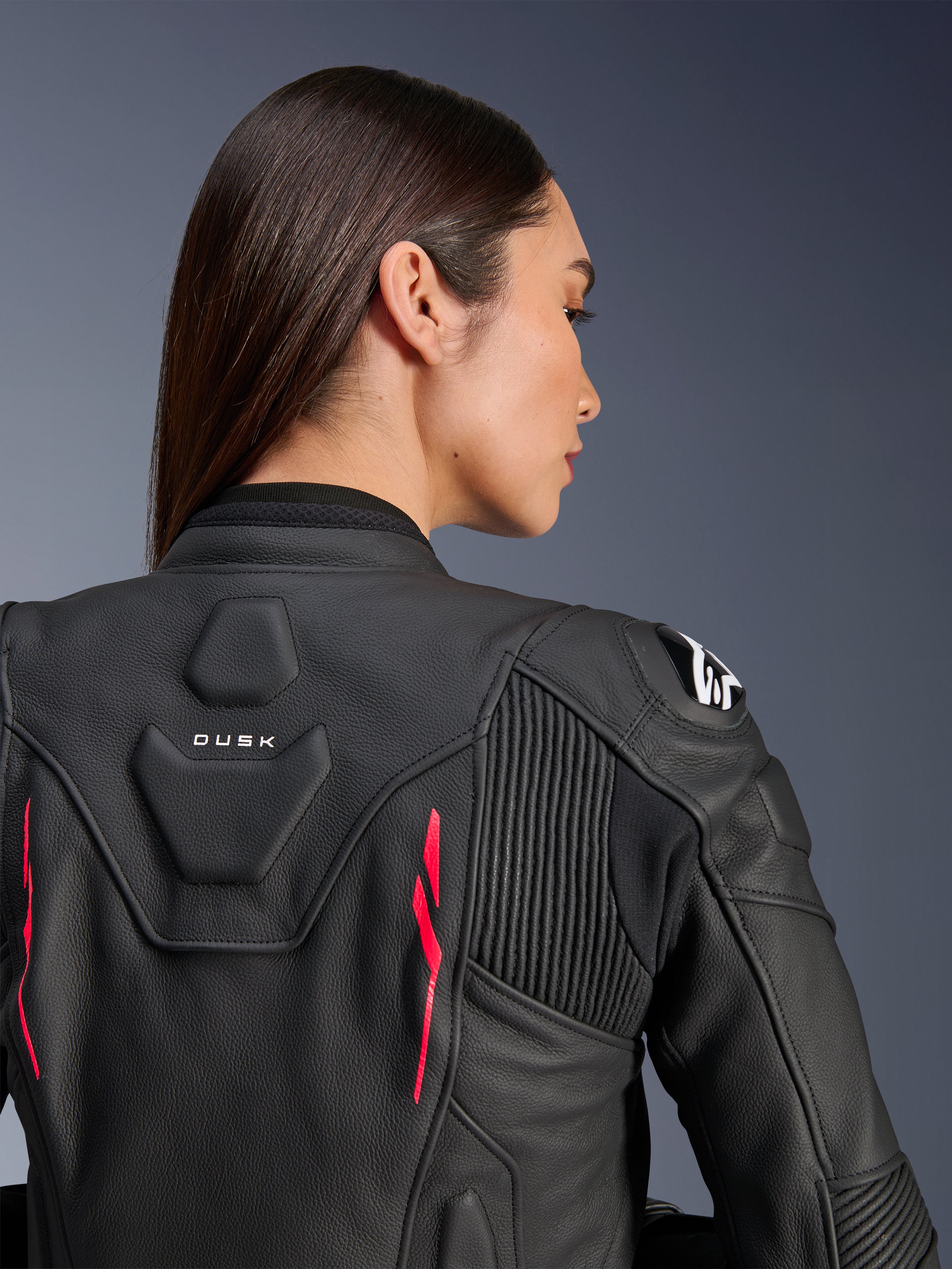 Vrouw draagt Alpinestars Stella Dusk leren jas, zwart leer met Diva Pink accenten en wit logo op de schouder, achteraanzicht met DUSK-branding en accordeon stretchpanelen, sportief ergonomisch ontwerp