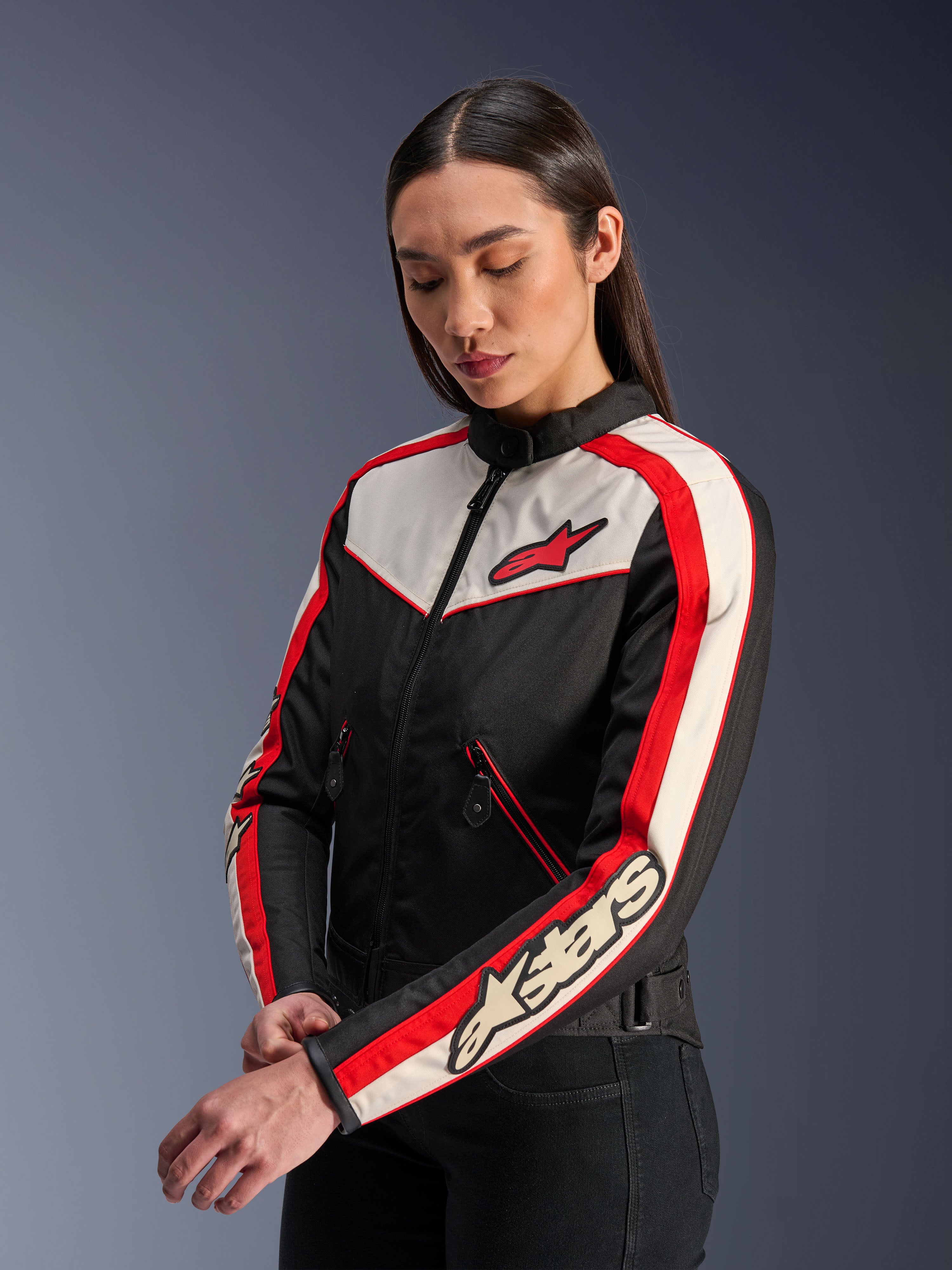 Alpinestars Stella T-Dyno WR Jas, textiele motorjas voor dames, zwart, ecru en felrood, vintage-geïnspireerd ontwerp met rood Alpinestars-logo op de borst en branding op de mouw, gedragen door een model in een studio-omgeving.