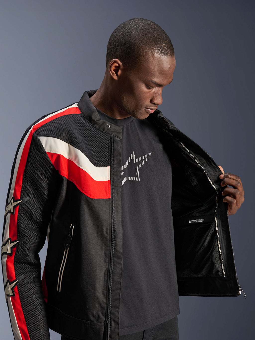 Alpinestars T-Dyno Air Jas, Zwart, Felrood en Ecru, mannelijk model opent de jas om de binnenvoering en het waterdichte compartiment te tonen, vintage-geïnspireerde textieljas met 3D-meshpanelen en sterlogo's, moderne motorrijuitrusting