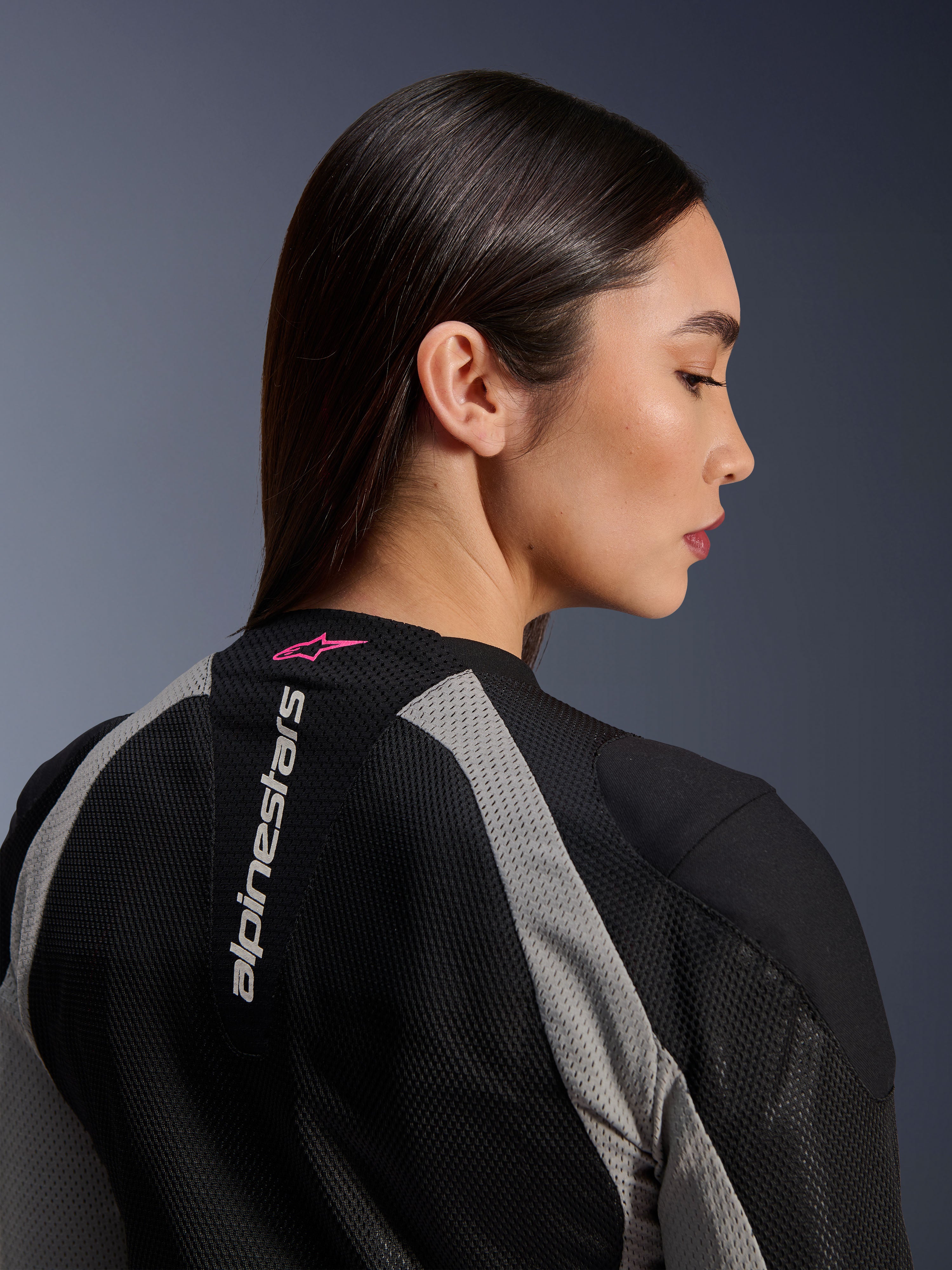 Close-up profiel van een vrouw die de Alpinestars Stella Troop-Air jas draagt, zwart en Ash Gray mesh-constructie, verticaal wit Alpinestars-logo op de bovenrug met een Pink Fluo ster-logo, lichte technische motoruitrusting voor dames