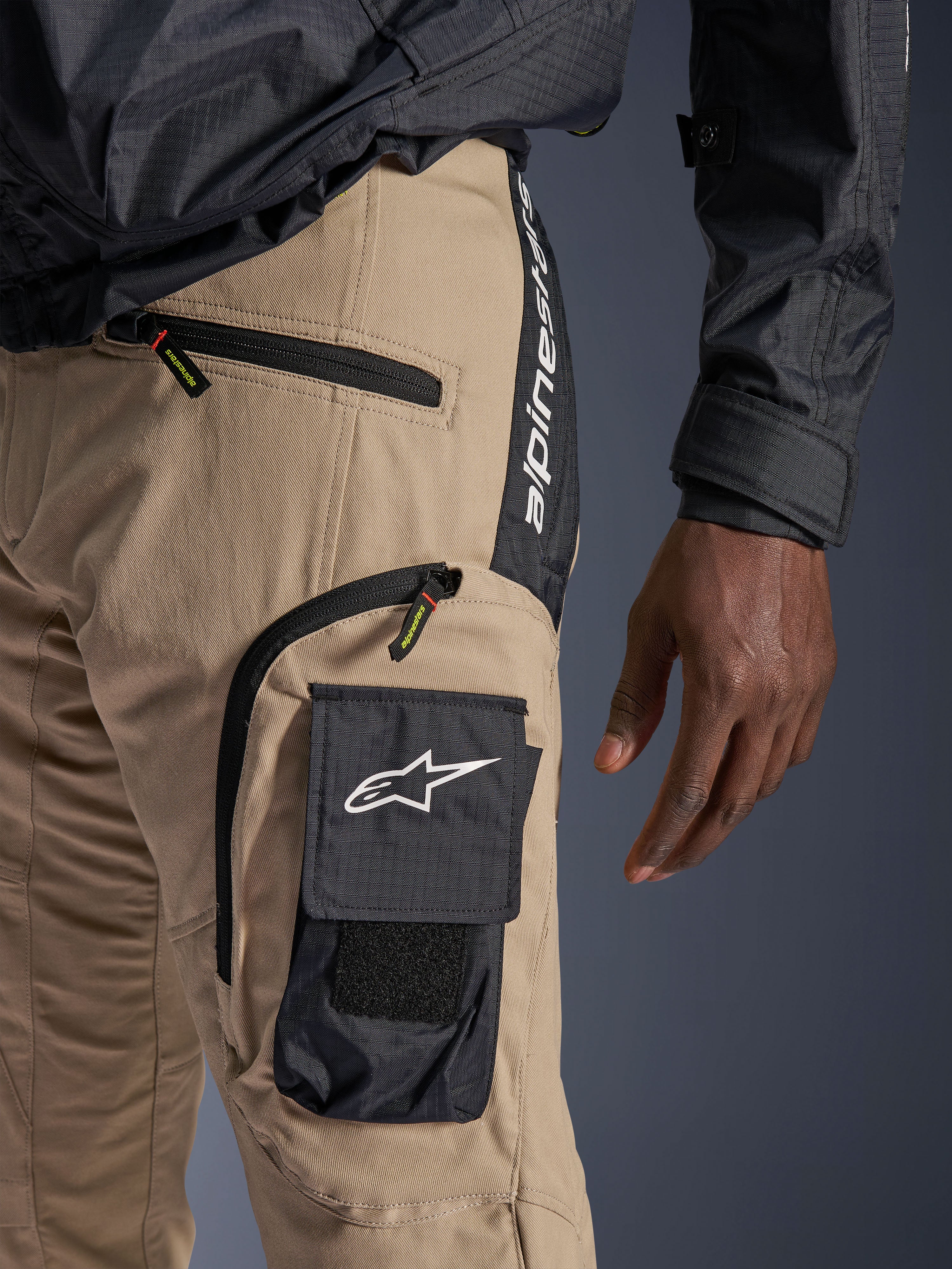 Alpinestars Acteon Broek in Sand, detailopname van de heup- en dijstreek, met een zwarte cargozak aan de zijkant met wit sterlogo, zakken met ritssluiting en verticale Alpinestars-branding op een zwart zijpaneel, urban motorkleding.