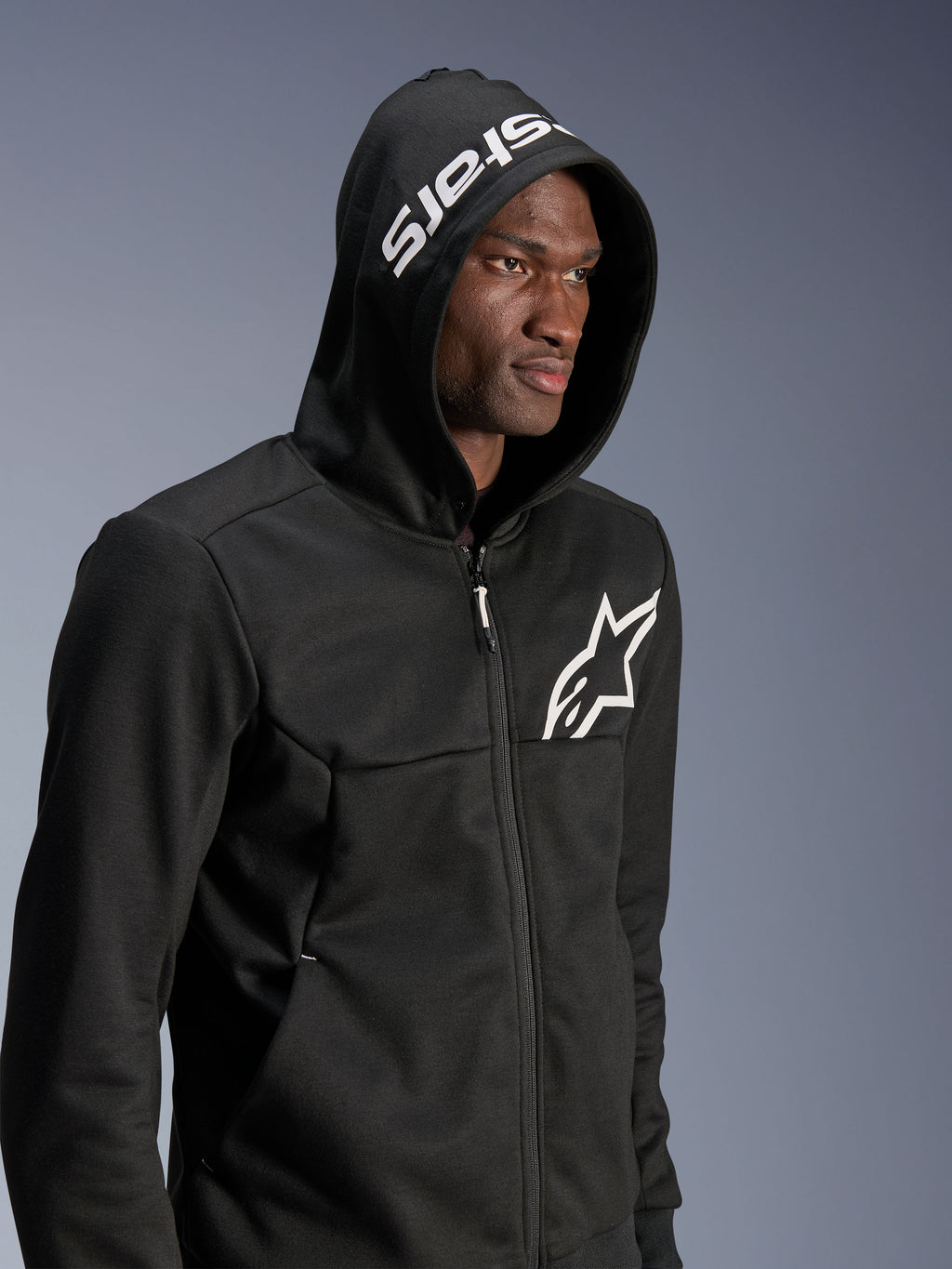 Alpinestars Chrome V2 Sport Hoodie, zwarte urban motorhoodie gedragen door model met de capuchon op, wit sterlogo op de borst en witte stars branding op de capuchon, tech fleece constructie, casual motor streetwear