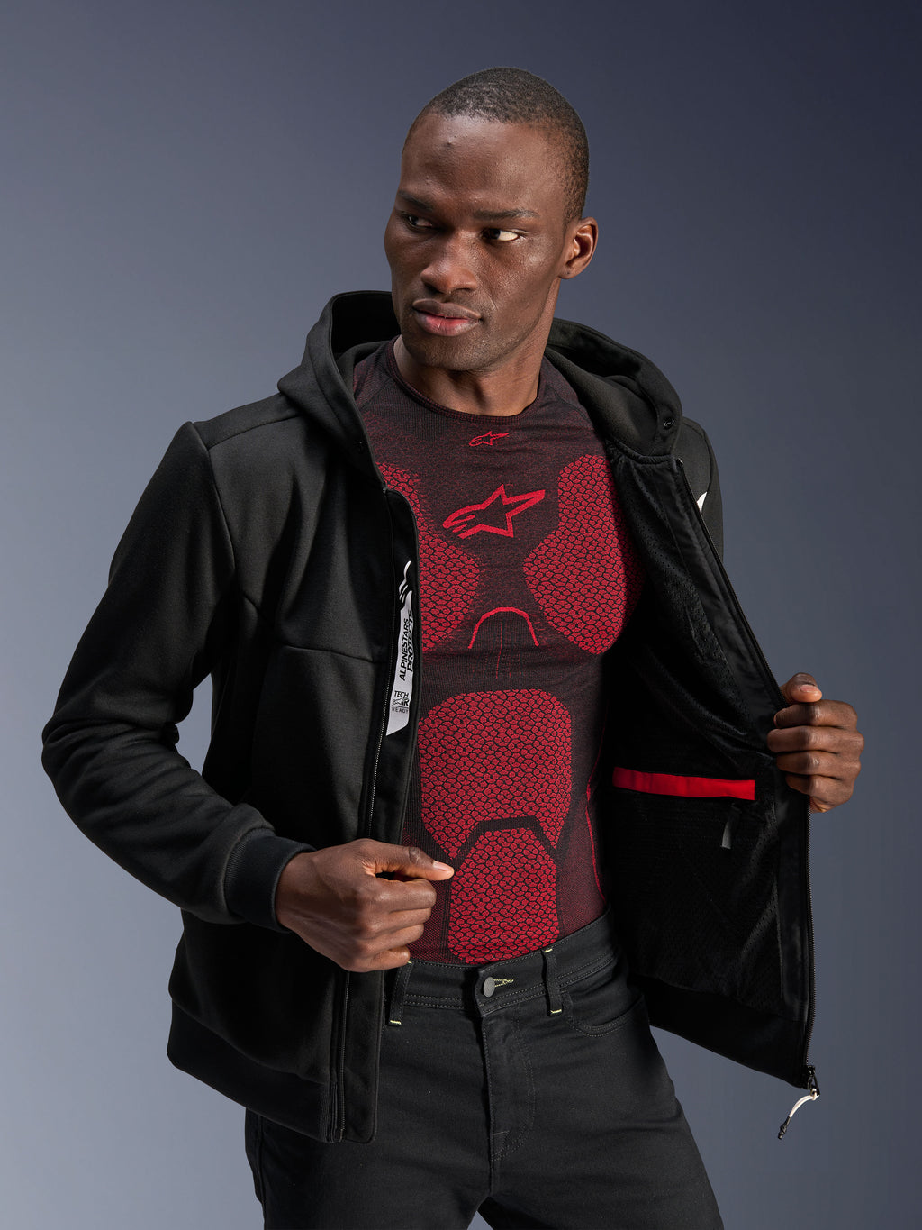 Alpinestars Chrome V2 Sport Hoodie, zwart, mannelijk model draagt de jas open om de binnenvoering, rode waterdichte zak en interne merkdetails te tonen, gedragen over een rood-zwarte basislaag, casual stedelijke motorkleding