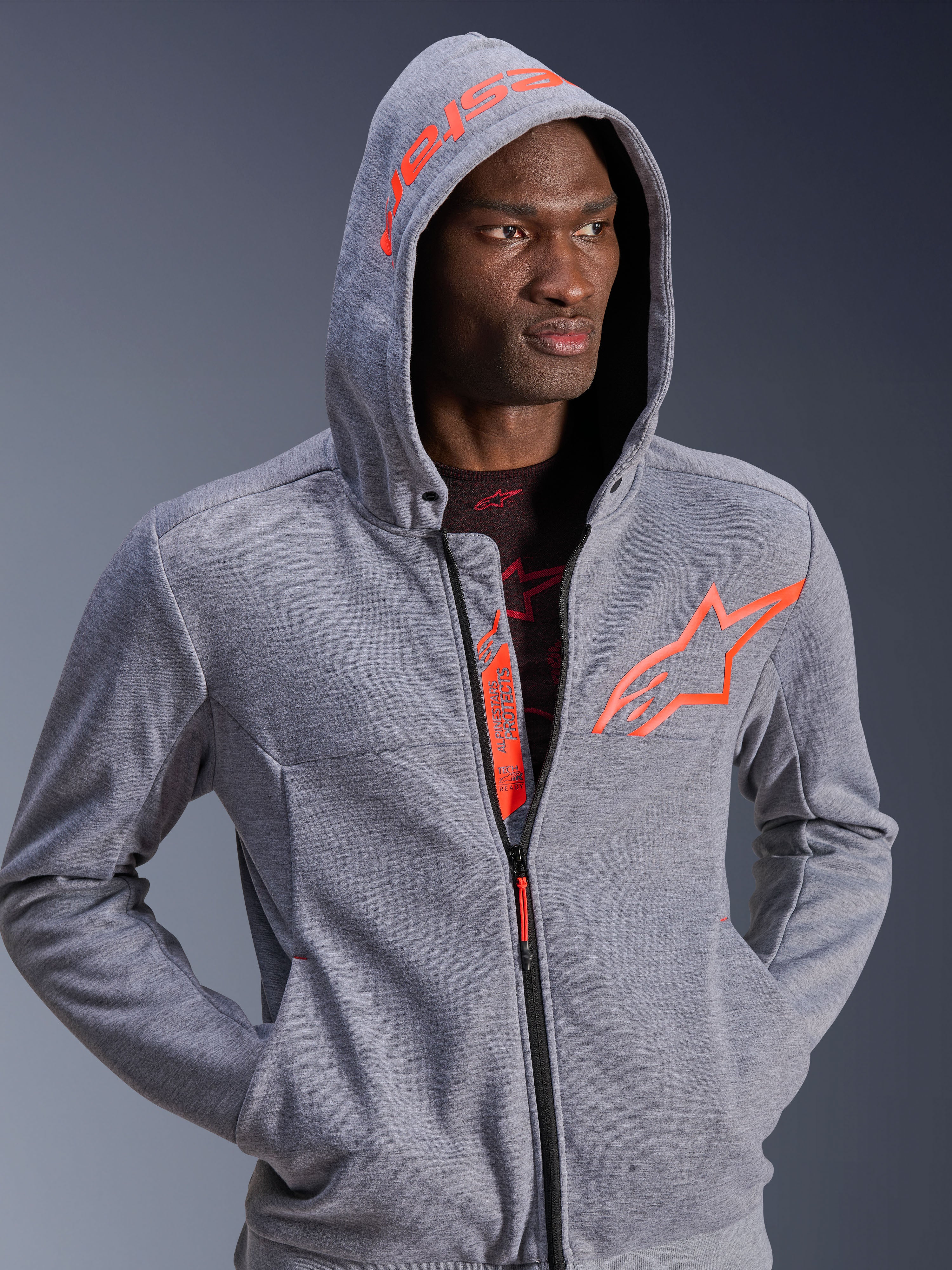 Alpinestars Chrome V2 Sport Hoodie, motorkleding, grijs gemêleerd en fluo rood, man met de capuchon op, voorzien van een groot oranje sterlogo op de borst en Alpinestars-tekst op de capuchon, casual urban ontwerp