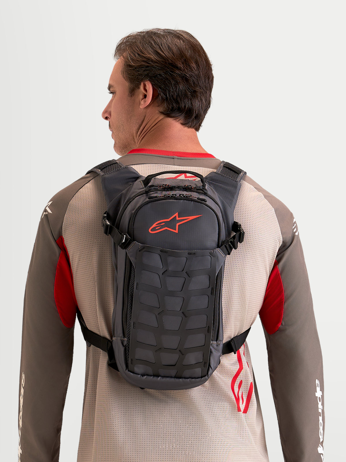 Alpinestars AT-9 rugzak, motocross rugzak gedragen door een model, walnoot- en donkergrijze textielconstructie met georganiseerde opbergvakken en een kenmerkend rood Astar-logo