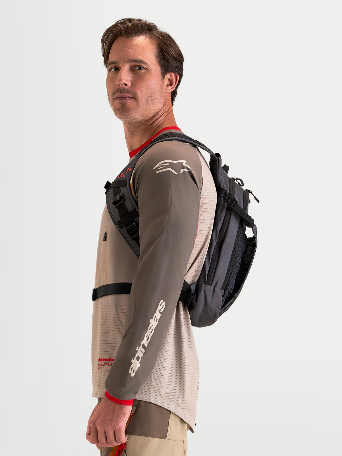Alpinestars AT-9 Rugzak, motocross rugzak in bruin en donkergrijs, gedragen door een mannelijk model met zicht op het zijharnas en de lichtgewicht textielconstructie