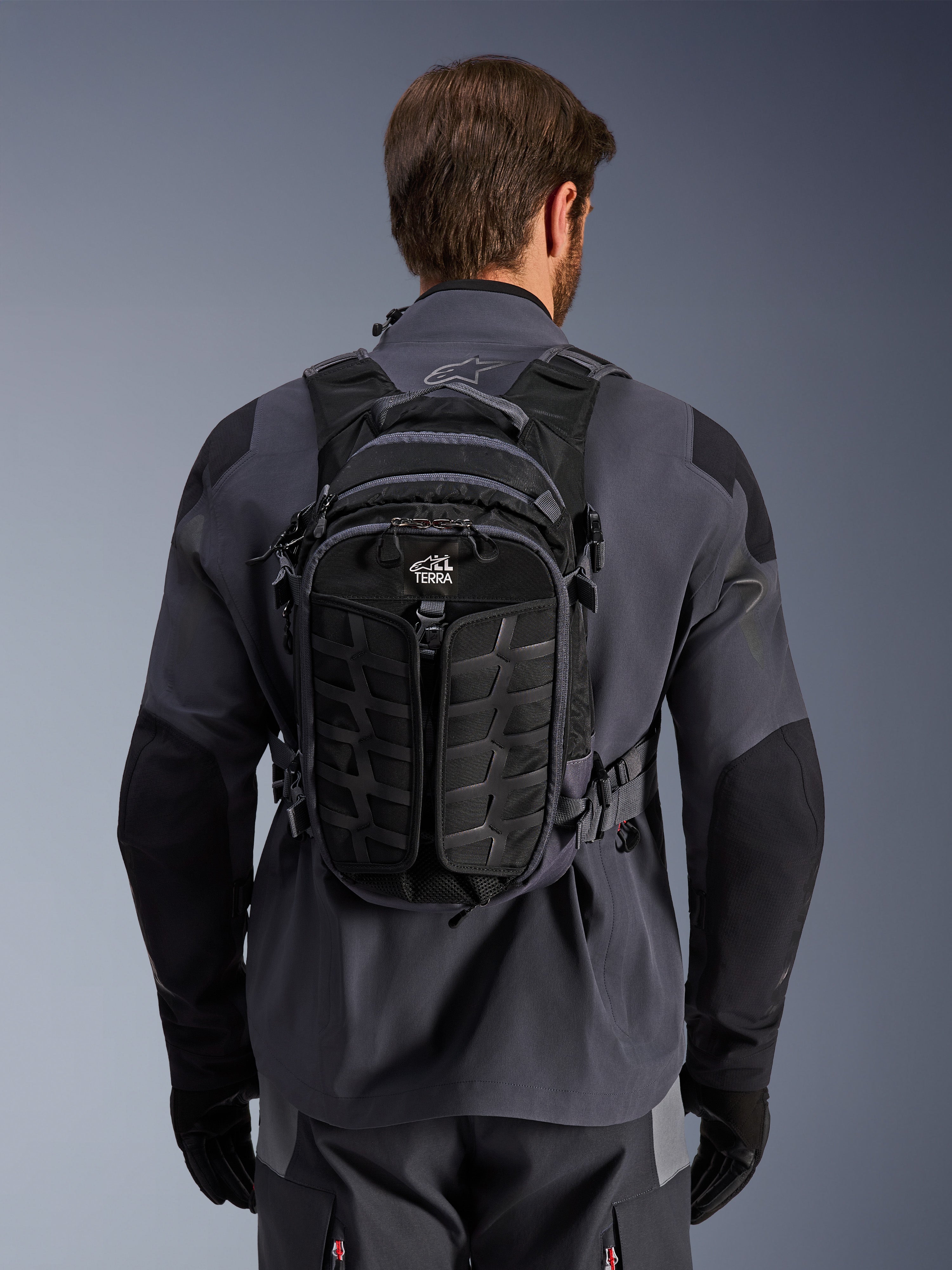 Alpinestars AT-15 Rugzak, adventure motorrugzak, Black Deep Gray, gedragen door een persoon over een grijs technisch motorjack, achteraanzicht met 'ALL TERRA' branding, ergonomisch ontwerp met versterkte panelen met textuur en stevige riemen.