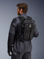 Alpinestars AT-15 rugzak, adventure motorrugzak, zwart en diepgrijs, gedragen door een rijder in een grijze jas, met een technisch ontwerp met MOLLE-webbing en 'TERRA'-branding, ergonomische off-road uitrusting