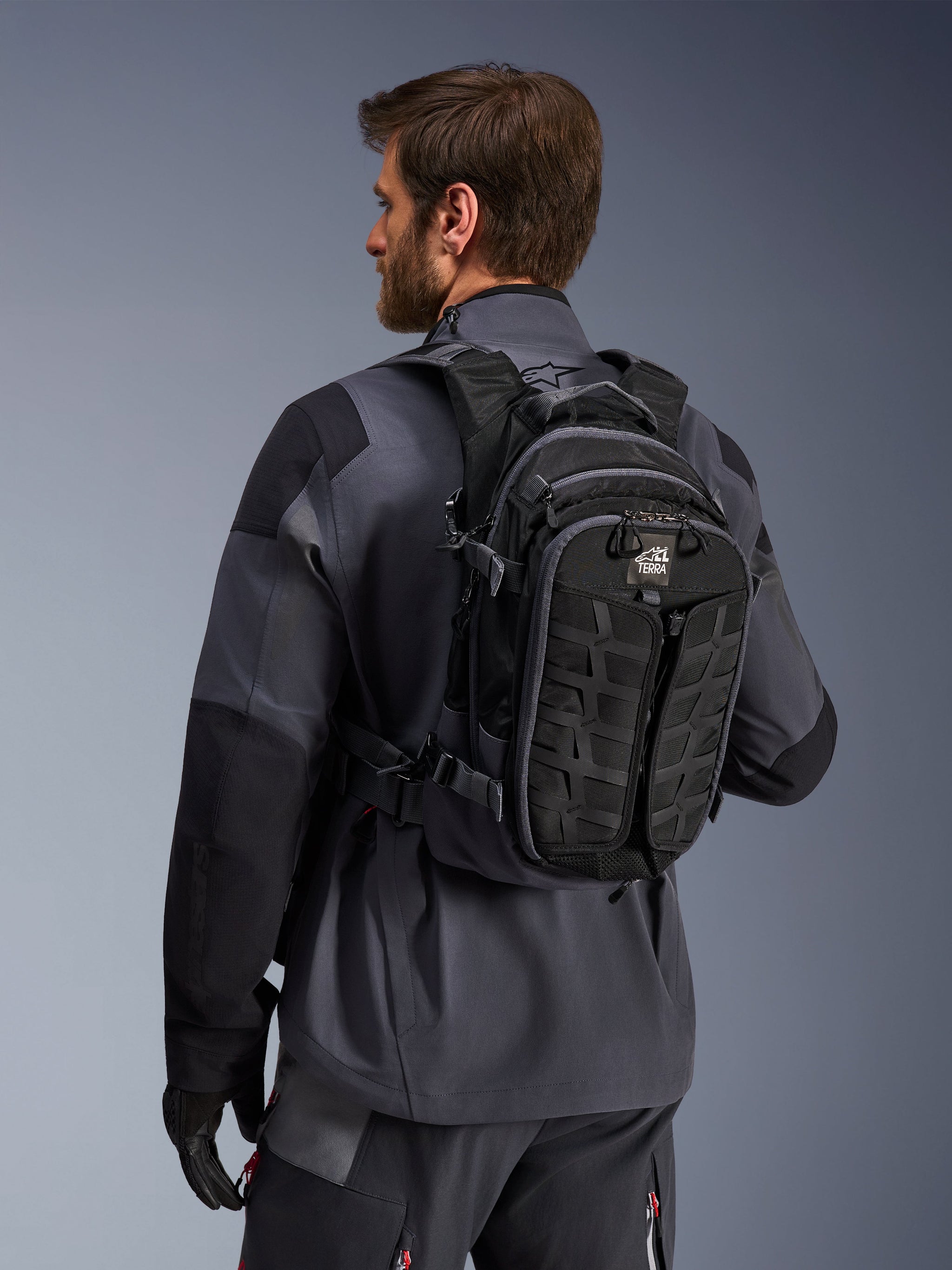 Alpinestars AT-15 rugzak, adventure motorrugzak, zwart en diepgrijs, gedragen door een rijder in een grijze jas, met een technisch ontwerp met MOLLE-webbing en 'TERRA'-branding, ergonomische off-road uitrusting