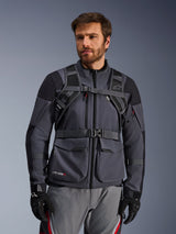 Alpinestars AT-15 rugzak draagsysteem, Black Deep Gray, vooraanzicht gedragen door een motorrijder over een grijze motorjas, voorzien van magnetische borstvergrendeling en stevige tailleriem, gecombineerd met zwarte handschoenen voor adventure motorrijden.