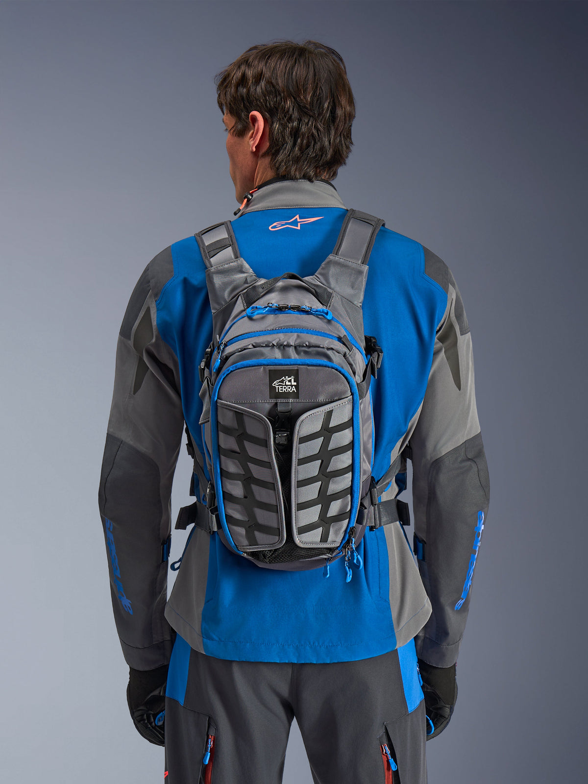 Alpinestars AT-15 rugzak, adventure motorrugzak, donkergrijs en diepgrijs, gedragen door een rijder over een blauw en grijs jack, met All Terra branding, ergonomisch structureel ontwerp, verstelbare banden en blauwe ritsaccenten.