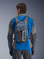 Alpinestars AT-15 rugzak, adventure motorrugzak, donkergrijs en diepgrijs, gedragen door een rijder over een blauw en grijs jack, met All Terra branding, ergonomisch structureel ontwerp, verstelbare banden en blauwe ritsaccenten.