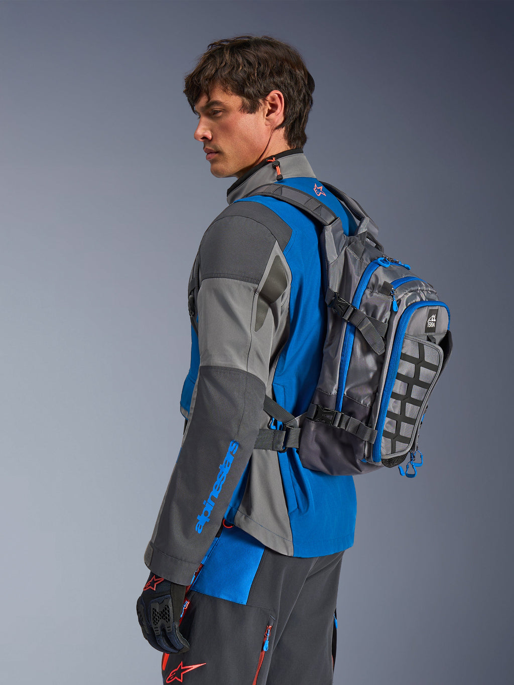 Alpinestars AT-15 rugzak, adventure motorrugzak, donkergrijs met blauwe accenten, gedragen door een rijder in bijpassende grijs-blauwe technische motorkleding, voorzien van MOLLE-paneel en robuuste constructie voor off-road avonturen.