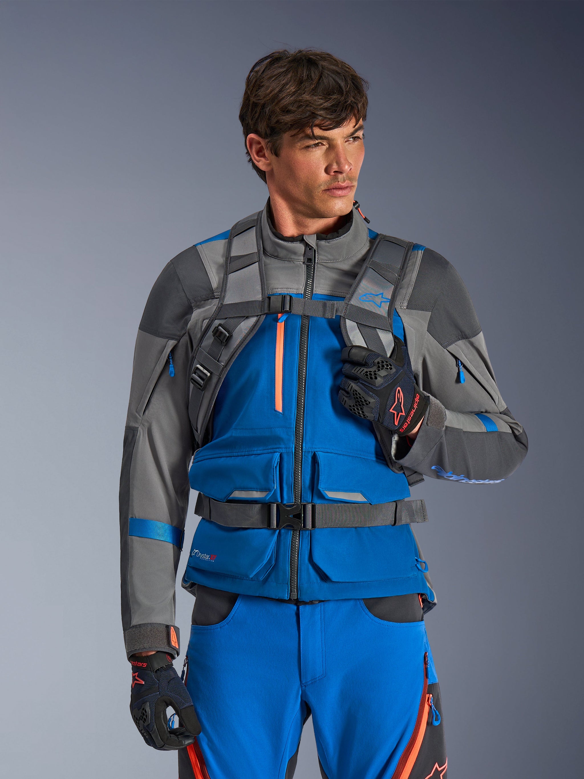 Alpinestars AT-15 rugzak, motorrugzak, donkergrijs, gedragen door een rijder in een blauwe en grijze adventure-jas en -broek, met technische grijze schouderbanden, magnetische borstsluiting en tailleriem, robuust ontwerp voor off-road rijden