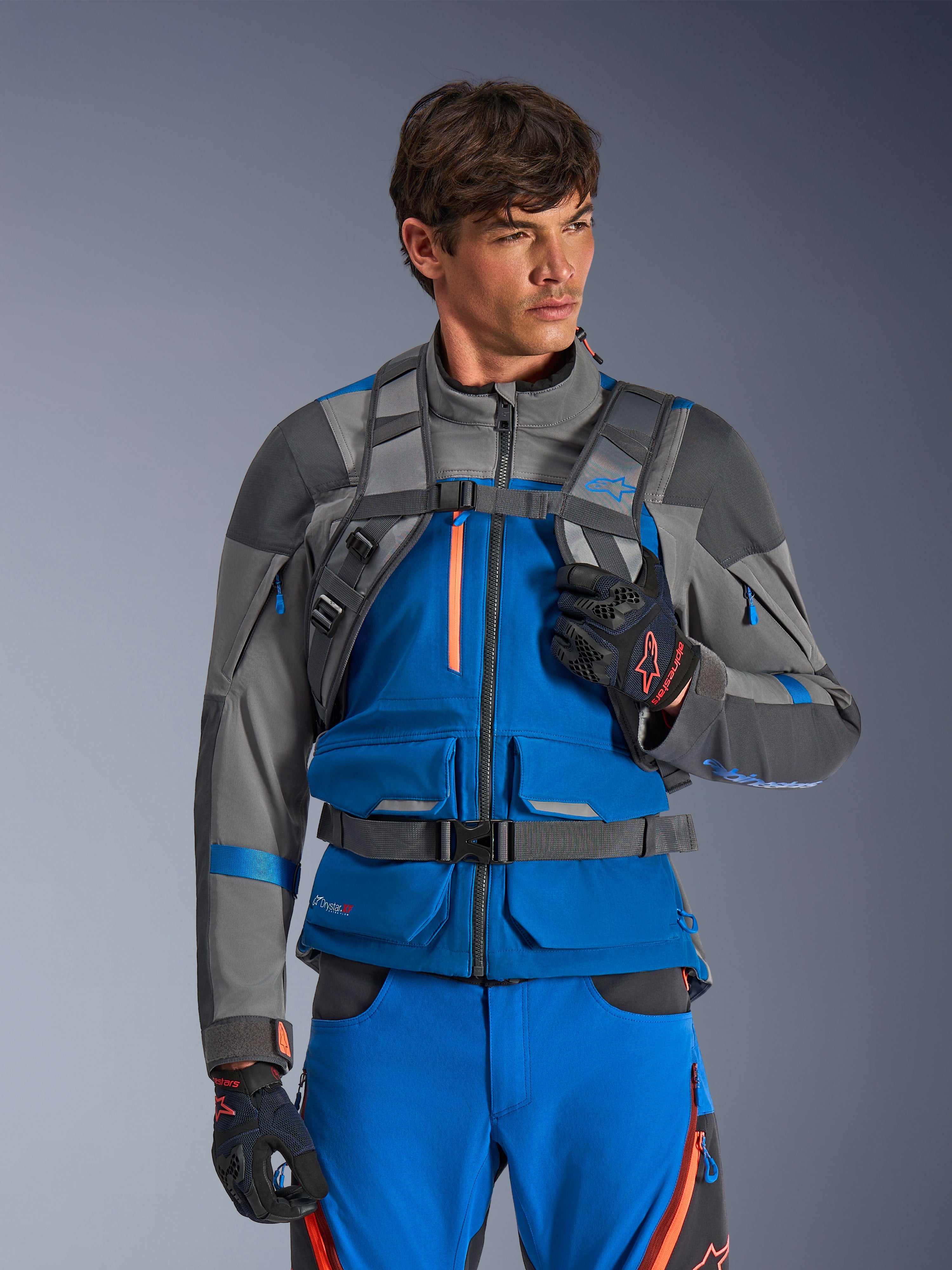Alpinestars AT-15 rugzak, motorrugzak, donkergrijs, gedragen door een rijder in een blauwe en grijze adventure-jas en -broek, met technische grijze schouderbanden, magnetische borstsluiting en tailleriem, robuust ontwerp voor off-road rijden