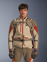 Adventure-rijder draagt de Alpinestars AT-15 Rugzak in Aluminum Walnut, vooraanzicht met borst- en taillebanden met magnetische sluiting, gecombineerd met een beige en rode adventure motorjas en -broek, technische off-road uitrusting