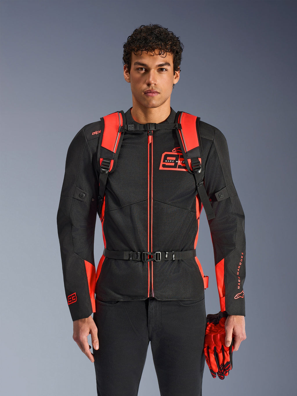 Alpinestars MM93 AMP3 rugzak, fluo rood en zwart, voorzien van verstelbare schouderbanden en borstriem met gesp, man draagt bijpassende MM93 motorjas en handschoenen, technische uitrusting uit de Marc Marquez-collectie