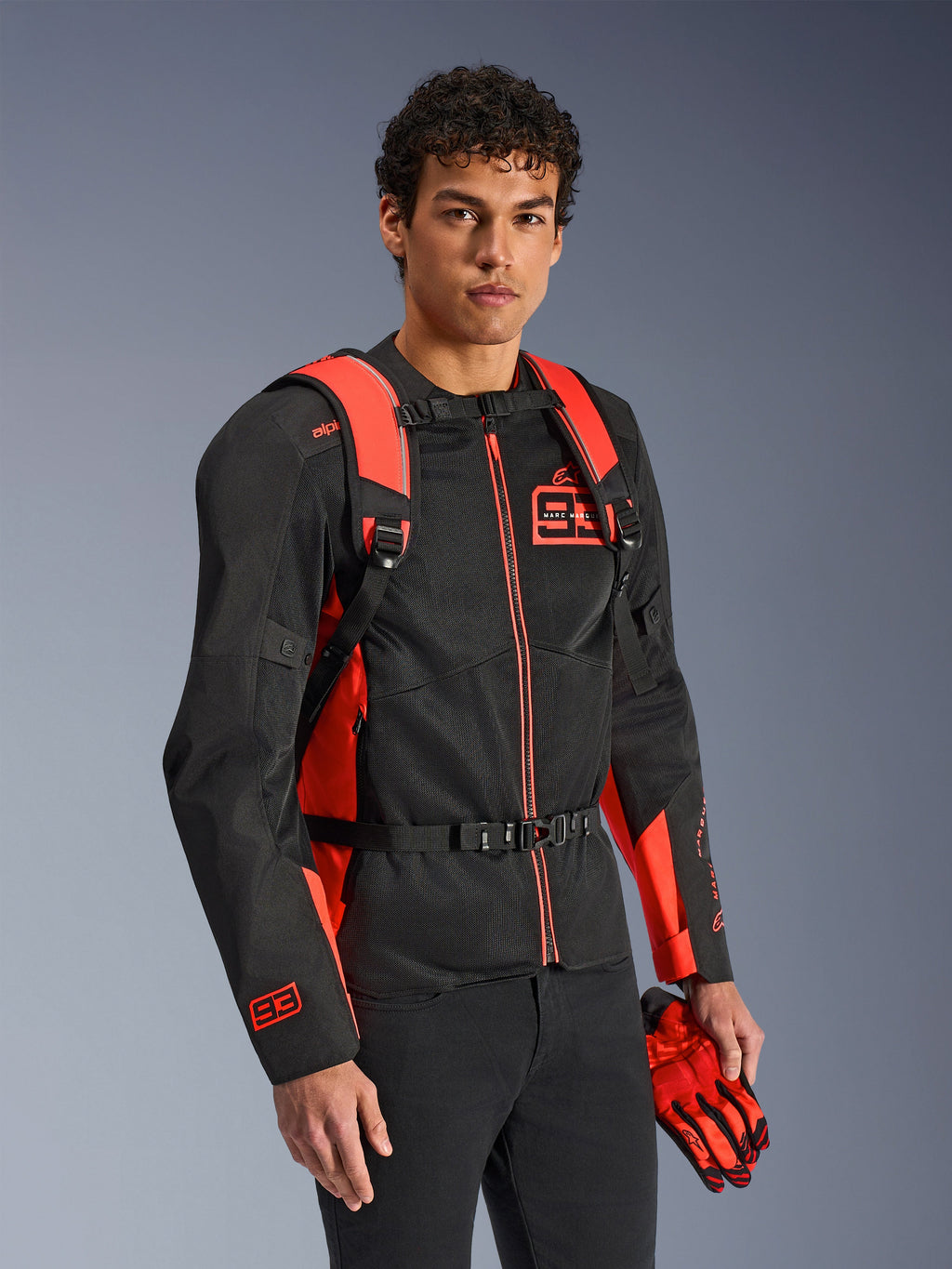 Alpinestars MM93 AMP3 rugzak, fluo rood en zwart, gedragen door een model met een bijpassende zwart-rode MM93 motorjas en handschoenen, vooraanzicht met nadruk op de verstelbare schouderbanden en borstsluiting, technische op racing geïnspireerde uitrusting.