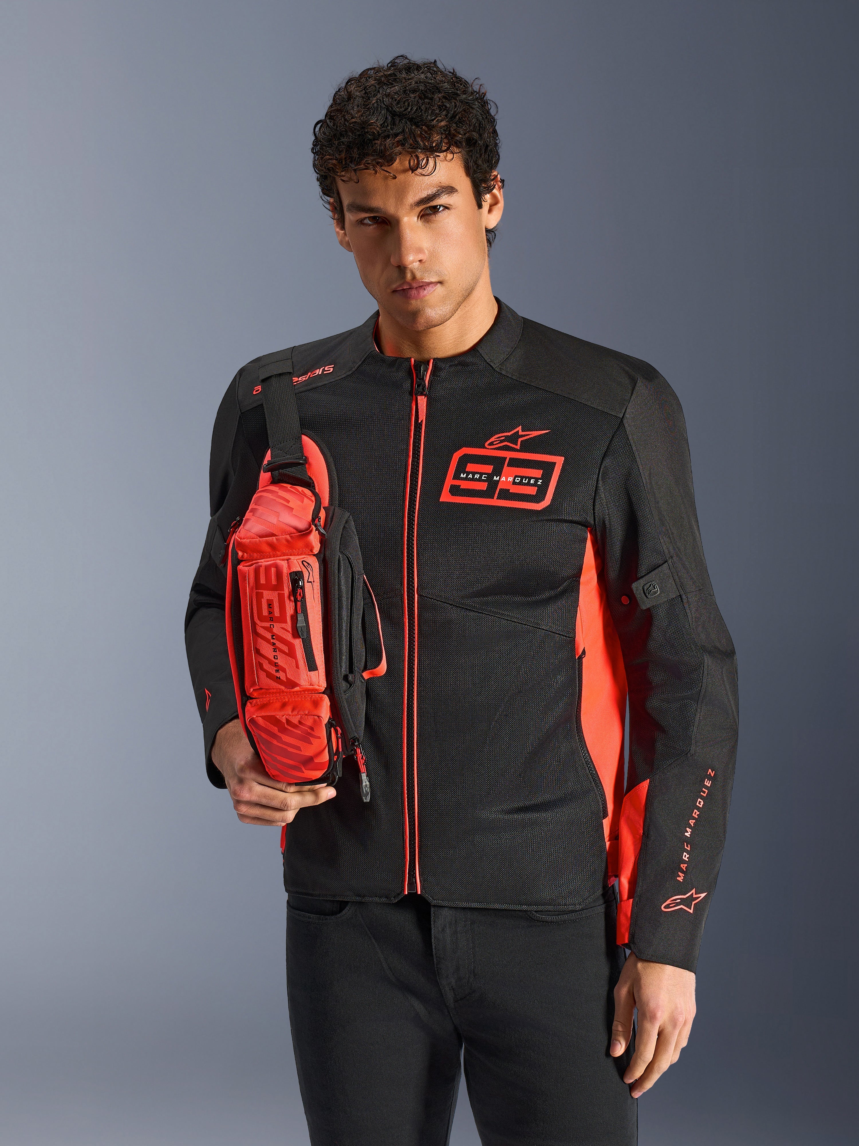 Alpinestars MM93 Waist Bag, heuptas gedragen als crossbodytas, fluo rood en zwart, man draagt bijpassend zwart en rood motorjack, voorzien van Marc Marquez 93-logo, sportief race-ontwerp