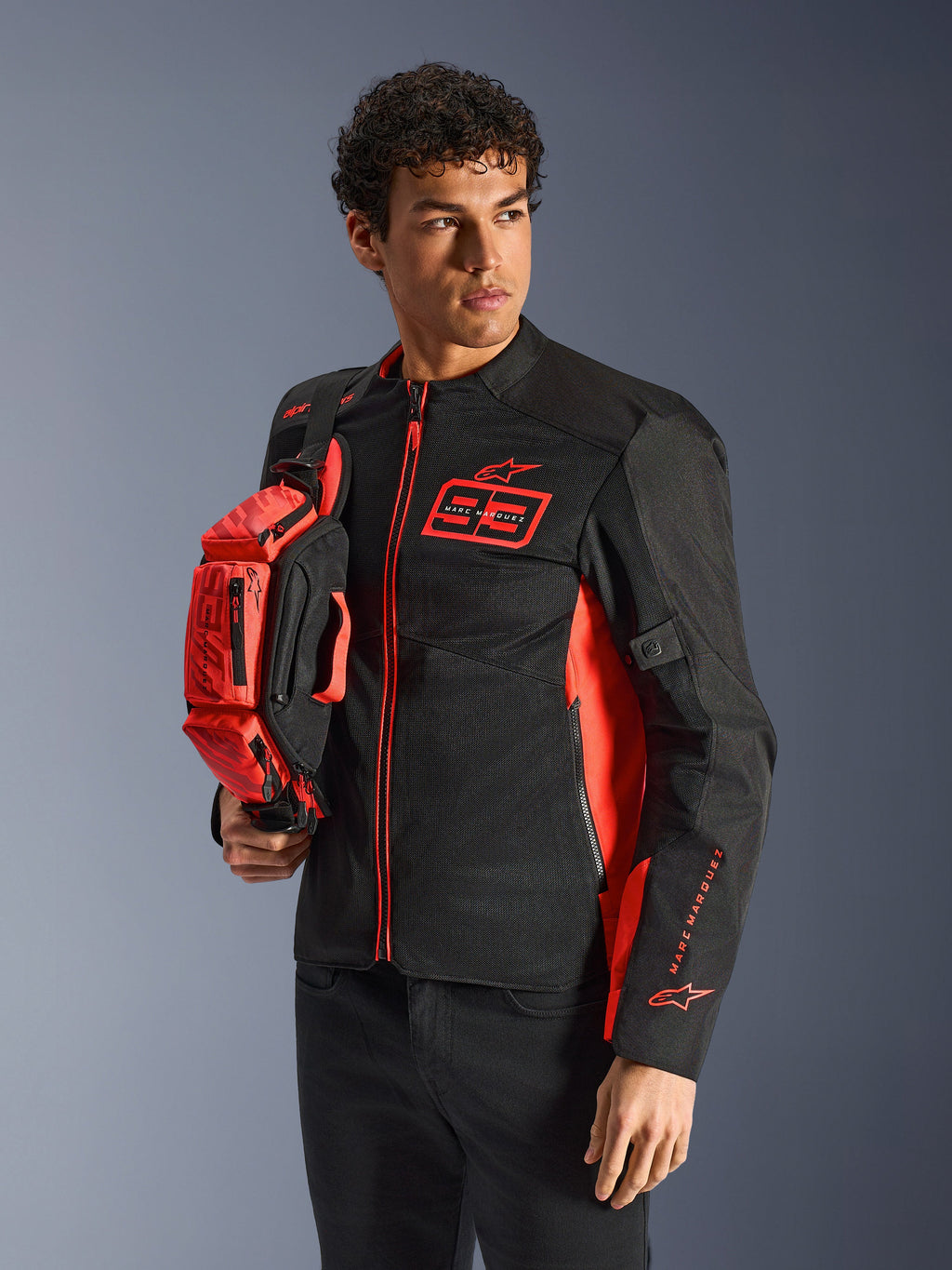Alpinestars MM93 heuptas, fluorescerend rood en zwart, vastgehouden door een model in een bijpassende zwart-rode motorjas, voorzien van meerdere vakken met ritssluiting en Marc Marquez-branding, praktische uitrusting voor sportief rijden
