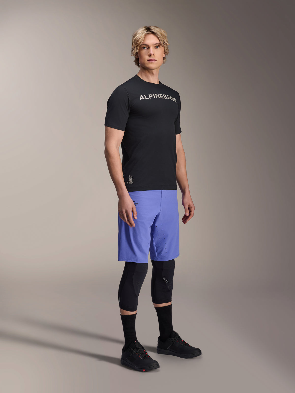 Mannelijk model draagt Alpinestars mountainbike-kleding, bestaande uit de zwarte A-Aria Switch SS Jersey en Baja Blue A-Aria Lite Shorts, gecombineerd met zwarte kniebeschermers, zwarte sokken en zwarte mountainbikeschoenen in een volledige studio-opname.