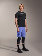 Mannelijk model draagt Alpinestars mountainbike-kleding, bestaande uit de zwarte A-Aria Switch SS Jersey en Baja Blue A-Aria Lite Shorts, gecombineerd met zwarte kniebeschermers, zwarte sokken en zwarte mountainbikeschoenen in een volledige studio-opname.