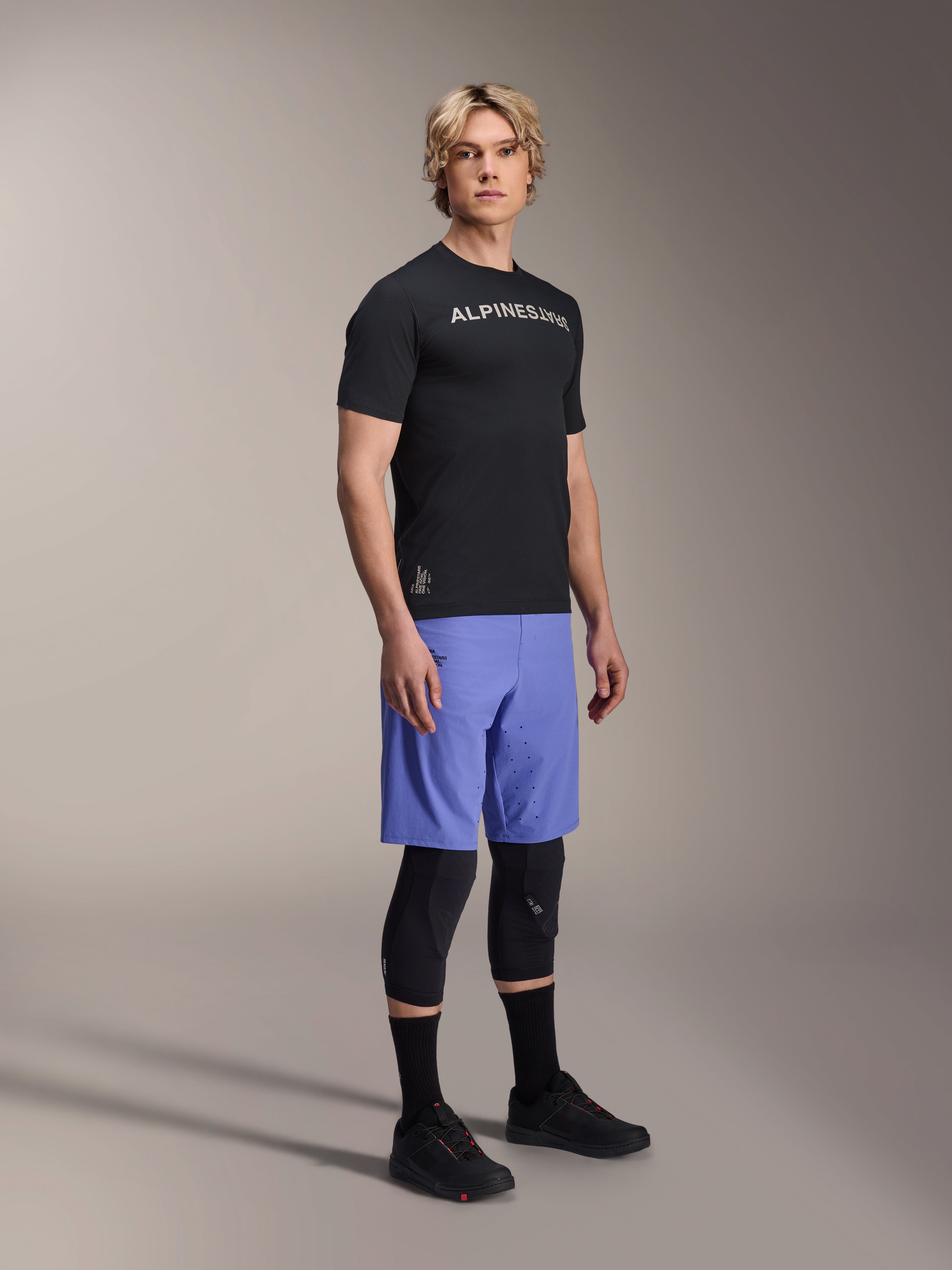Mannelijk model draagt Alpinestars mountainbike-kleding, bestaande uit de zwarte A-Aria Switch SS Jersey en Baja Blue A-Aria Lite Shorts, gecombineerd met zwarte kniebeschermers, zwarte sokken en zwarte mountainbikeschoenen in een volledige studio-opname.